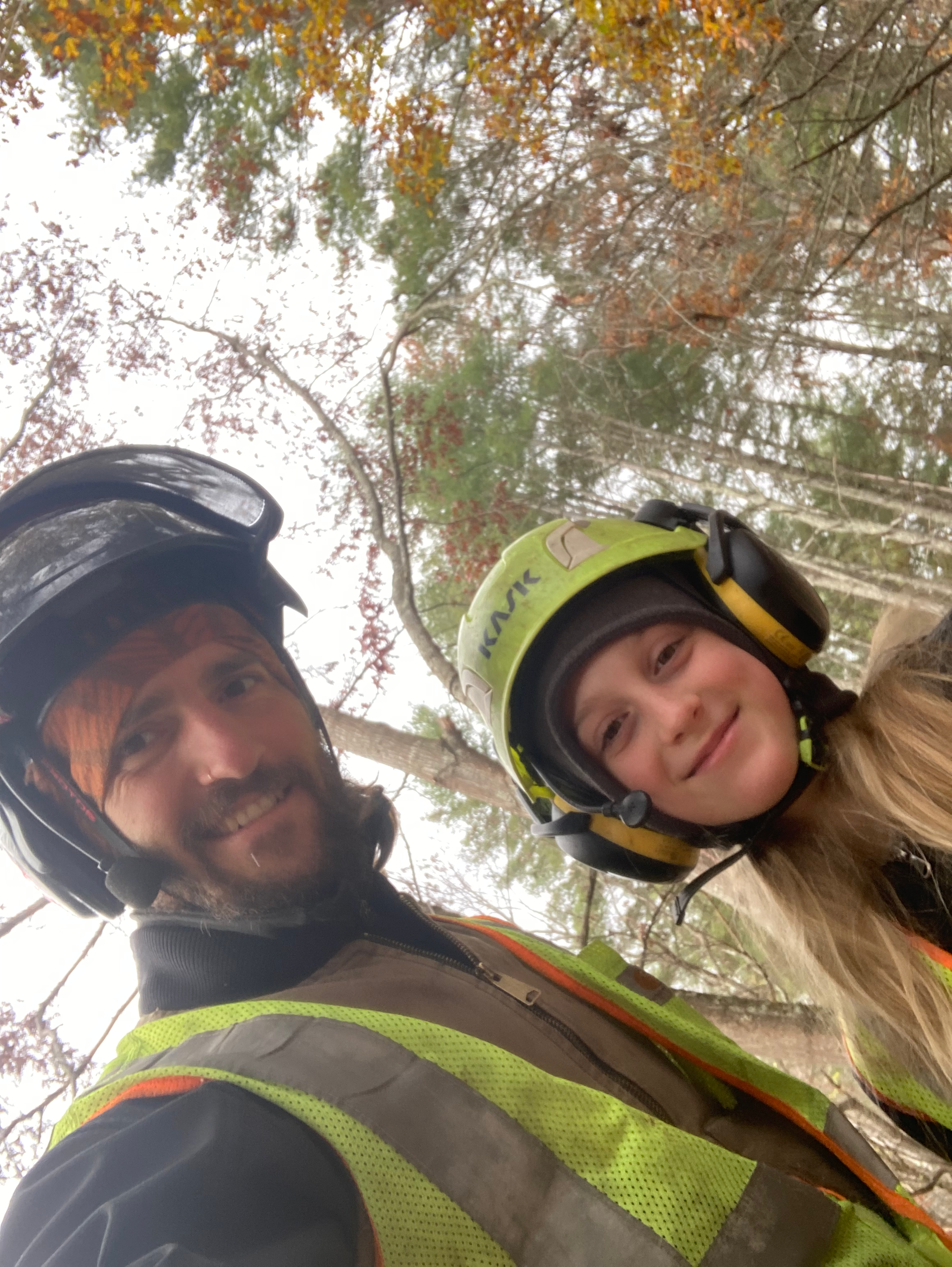 A man and his daughter wearing a safety helmet and a high-visibility vest with reflective stripes, standing outdoors among trees with autumn-colored leaves. Woodwitch Arborists, Tree Preservation & Removal in WNC