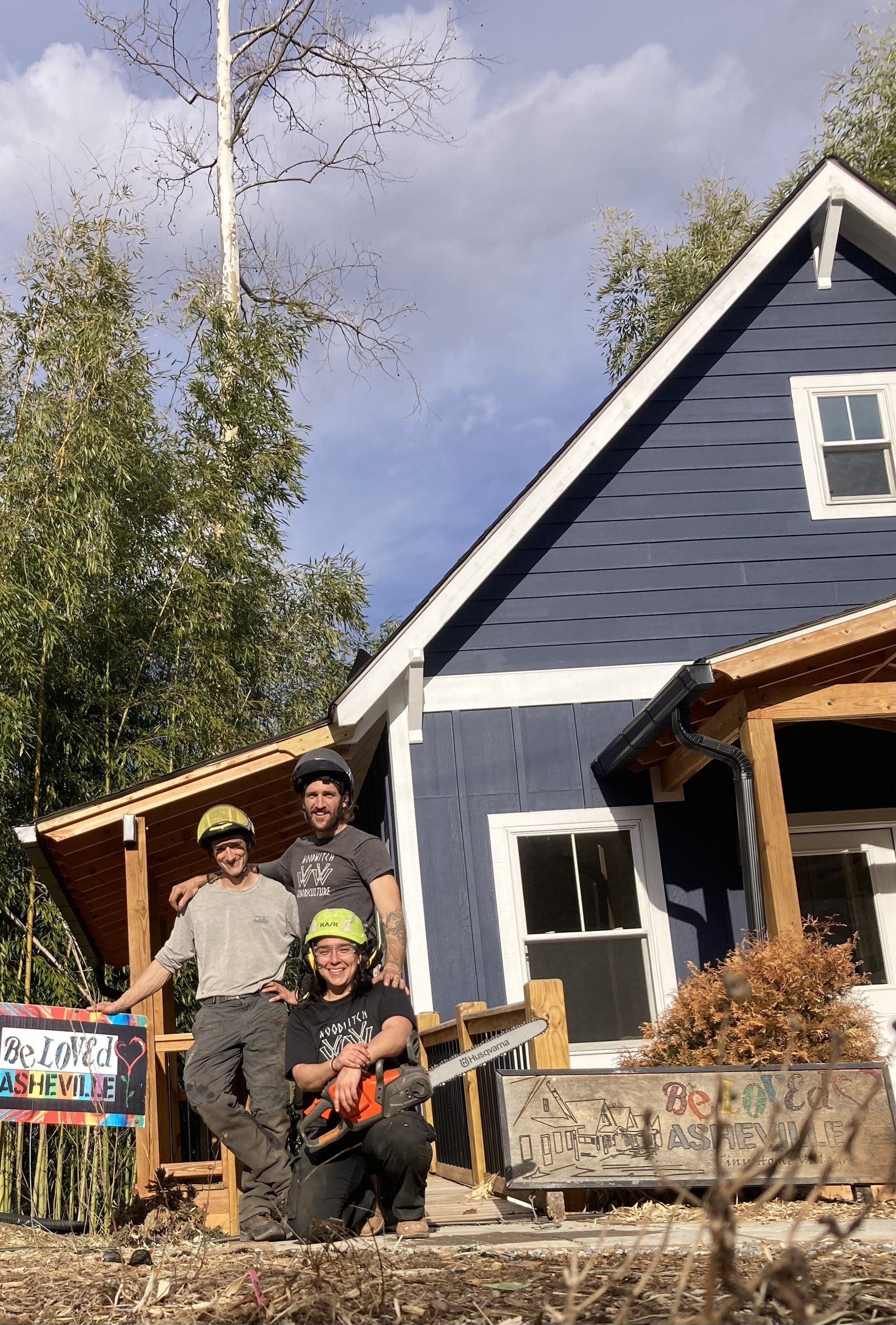 People in safety helmets and casual clothing standing on a porch in front of a house with blue siding and white trim. The porch has a wooden railing and a sign that reads 'Be Loved Asheville.' Trees with some leaves are in the background, and the sky is partly cloudy.