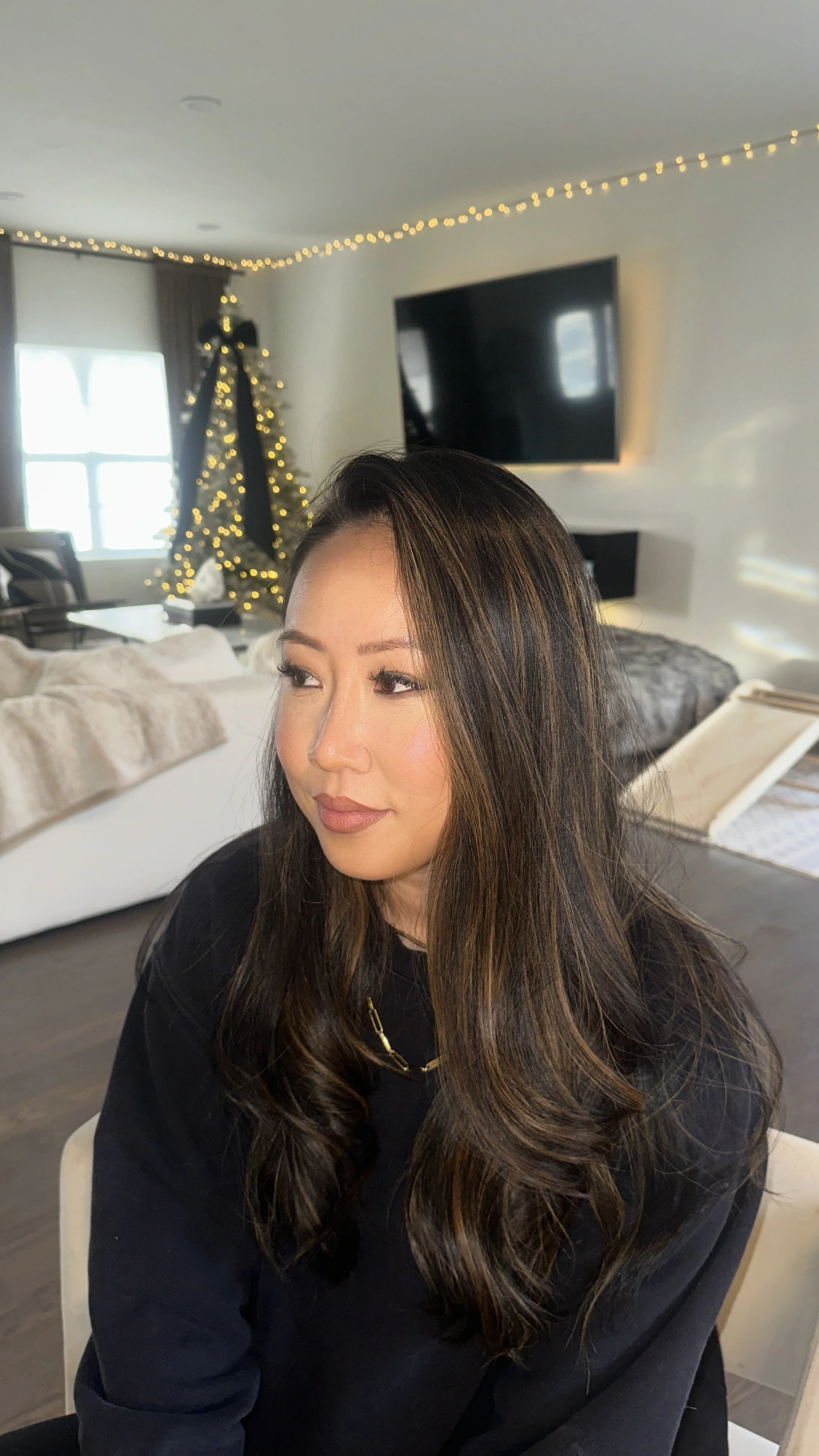 A woman with long, dark, wavy hair and light makeup sitting indoors with a Christmas tree decorated with lights and a large black ribbon in the background.