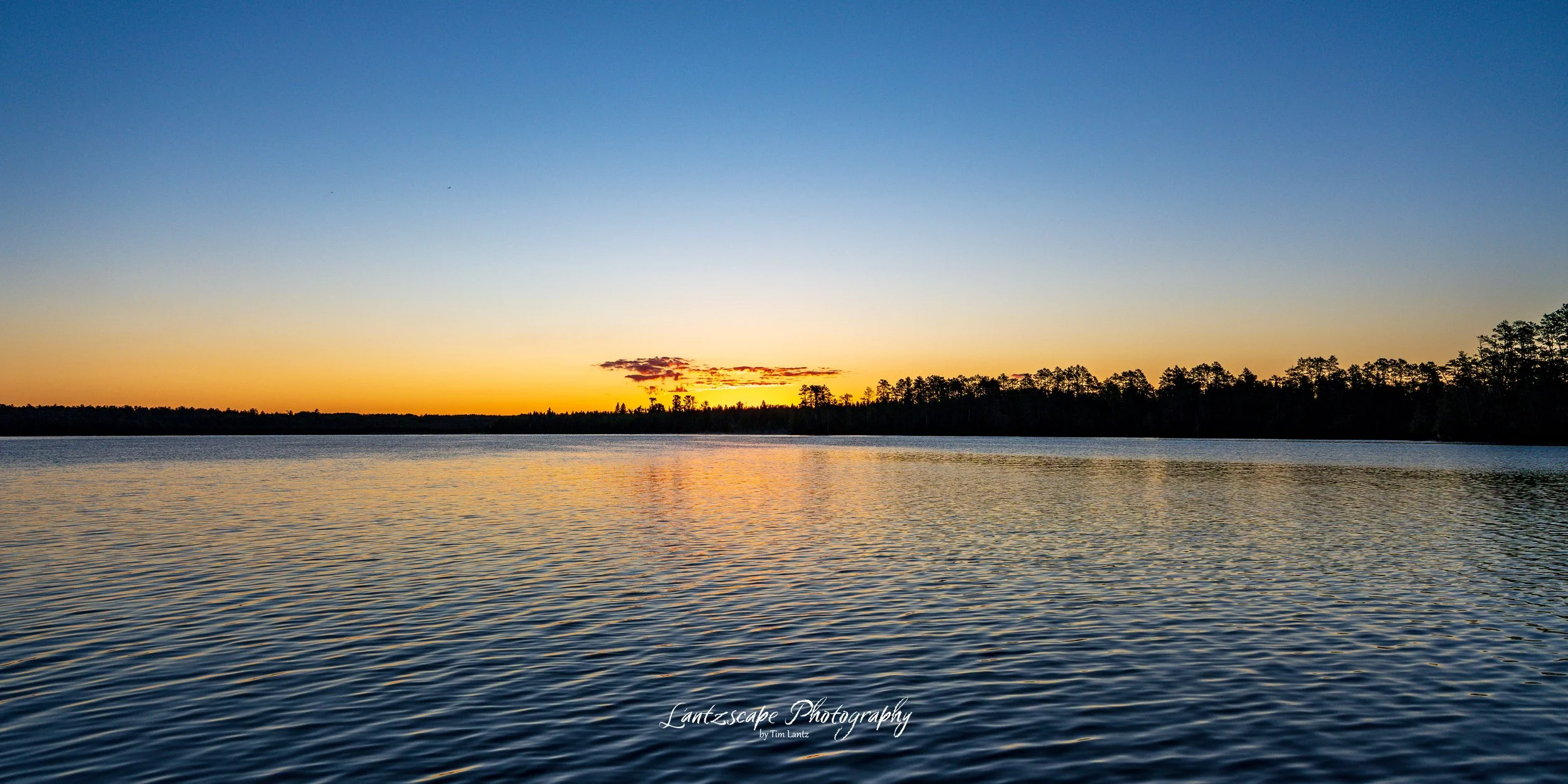 A peaceful lake at sunset with a calm water surface, a silhouette of trees on the horizon, and a clear sky transitioning from blue to orange near the horizon.