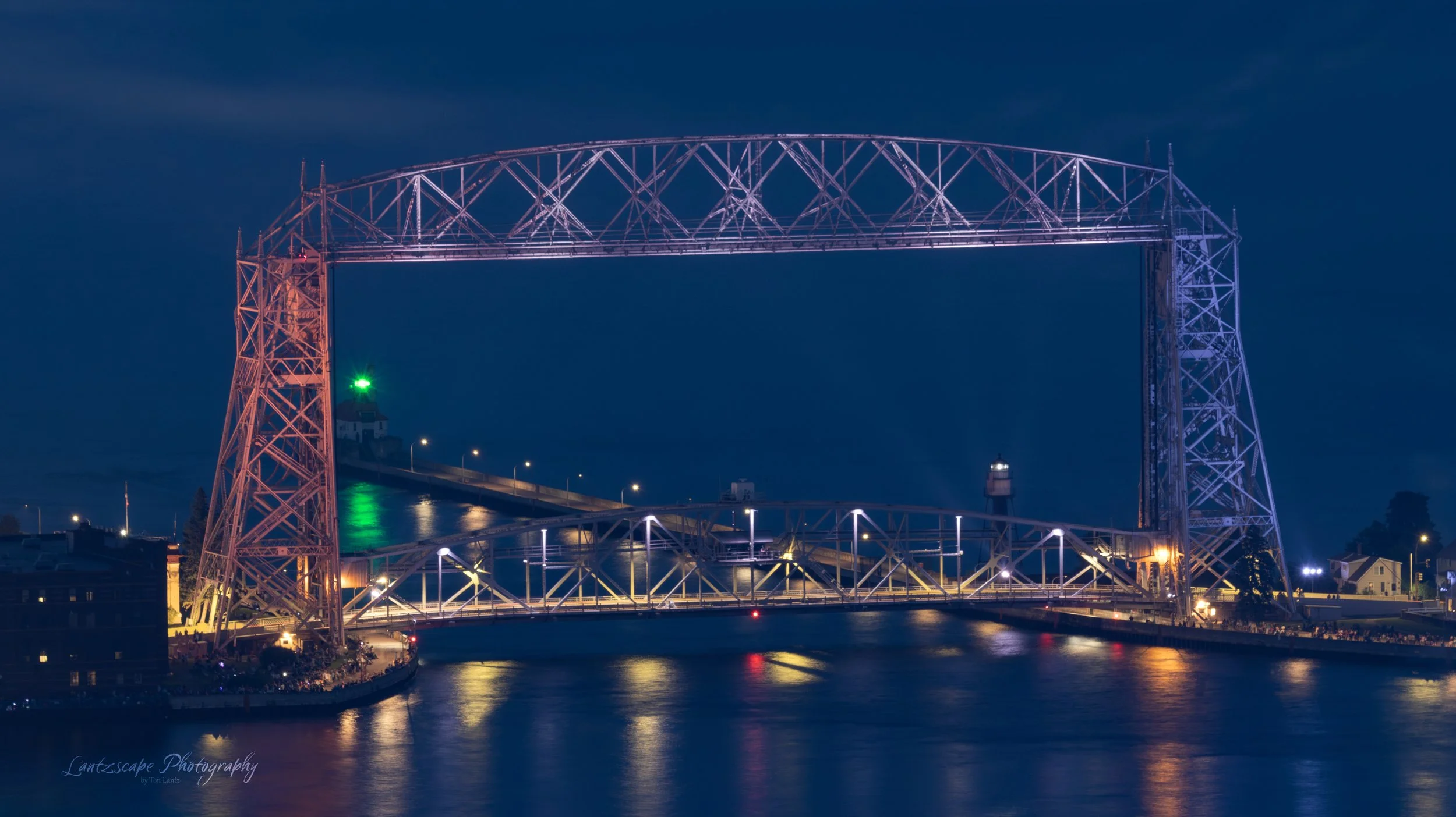 Nighttime view of the Hennepin Avenue Bridge in Minneapolis, illuminated with purple and white lights, reflecting on the river.