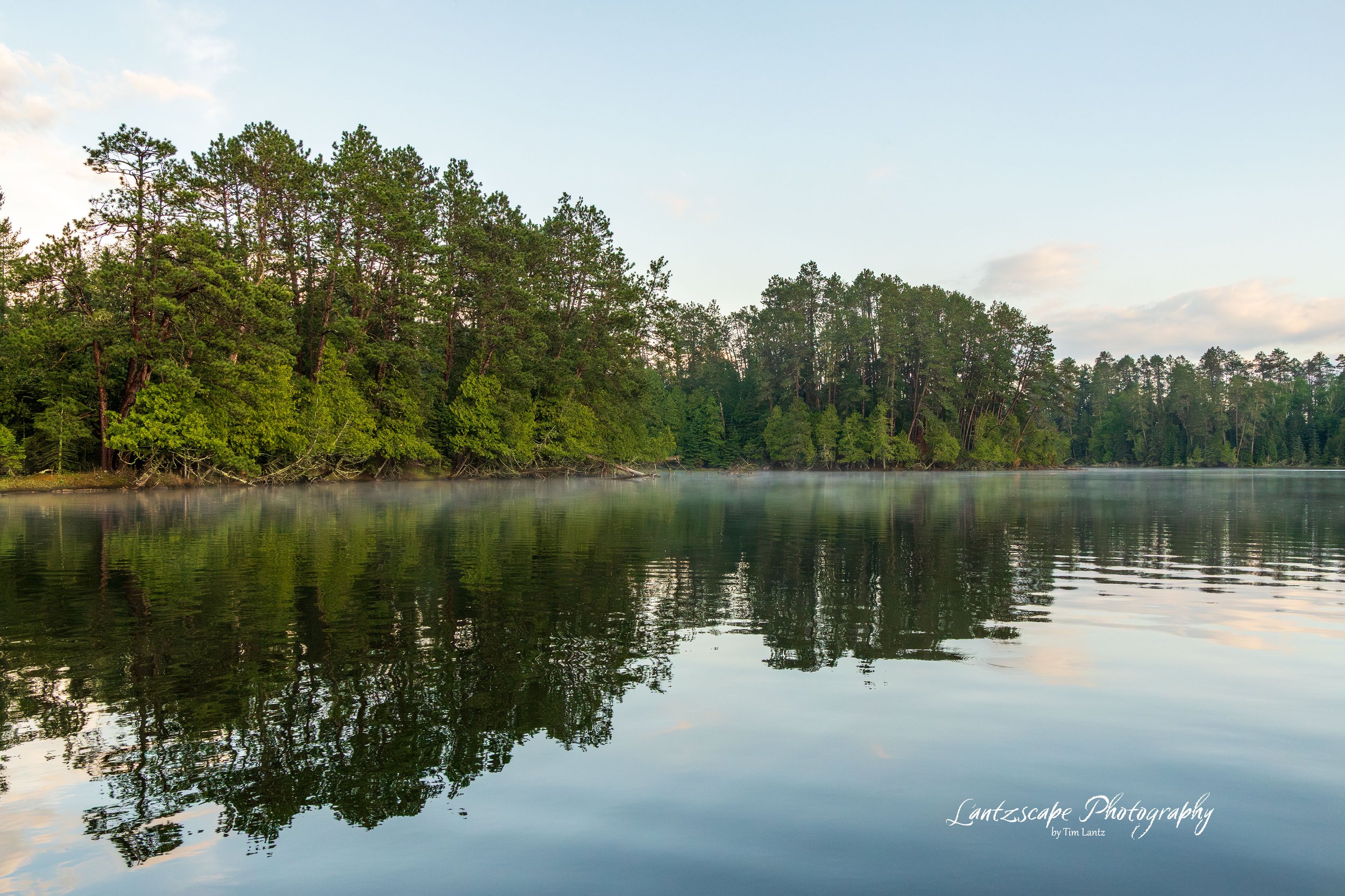 Calm lake with glassy water reflecting a forested shoreline of green trees, under a partly cloudy sky.