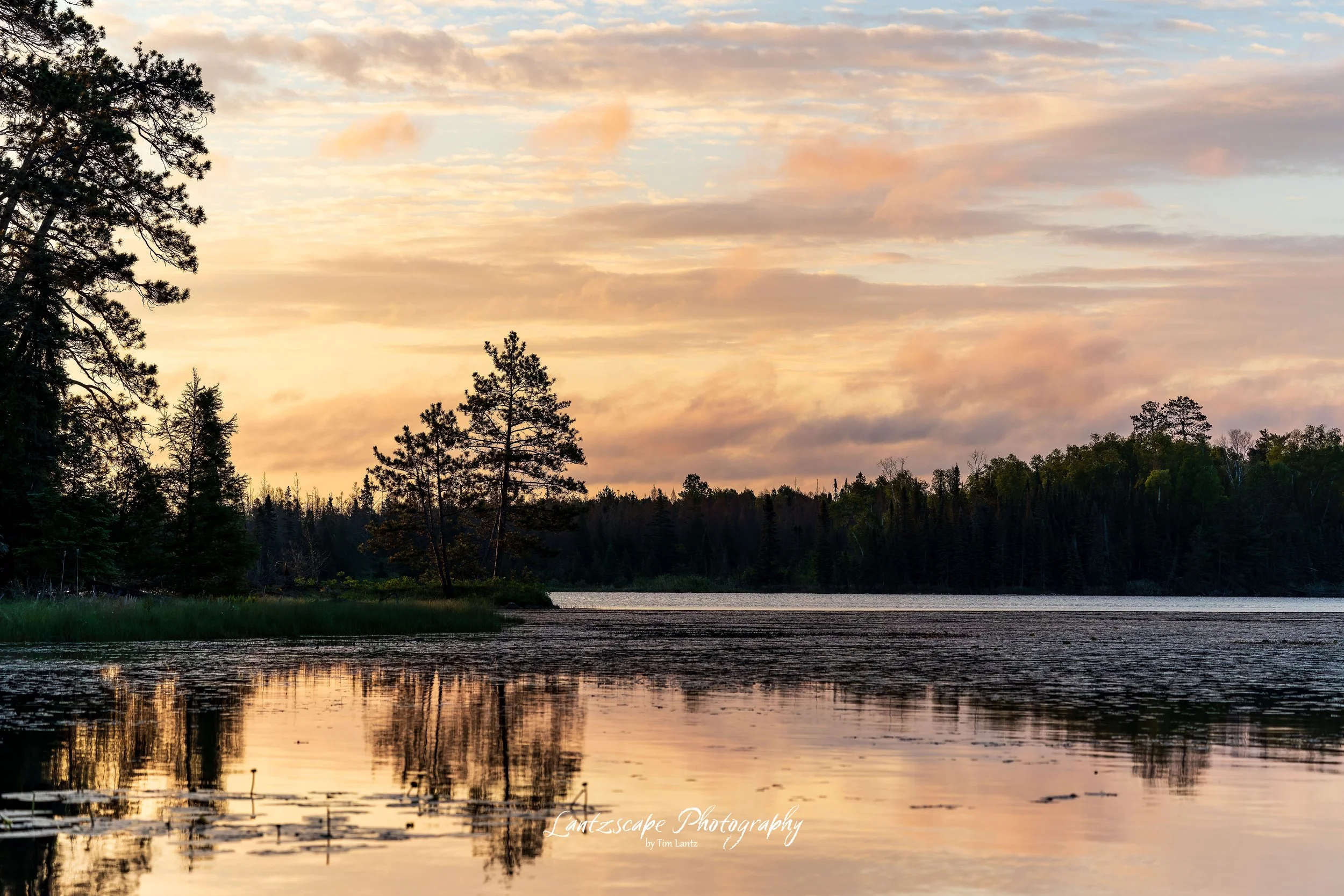 A sunset over a calm lake with trees along the shoreline and a reflection of the sky and trees on the water.