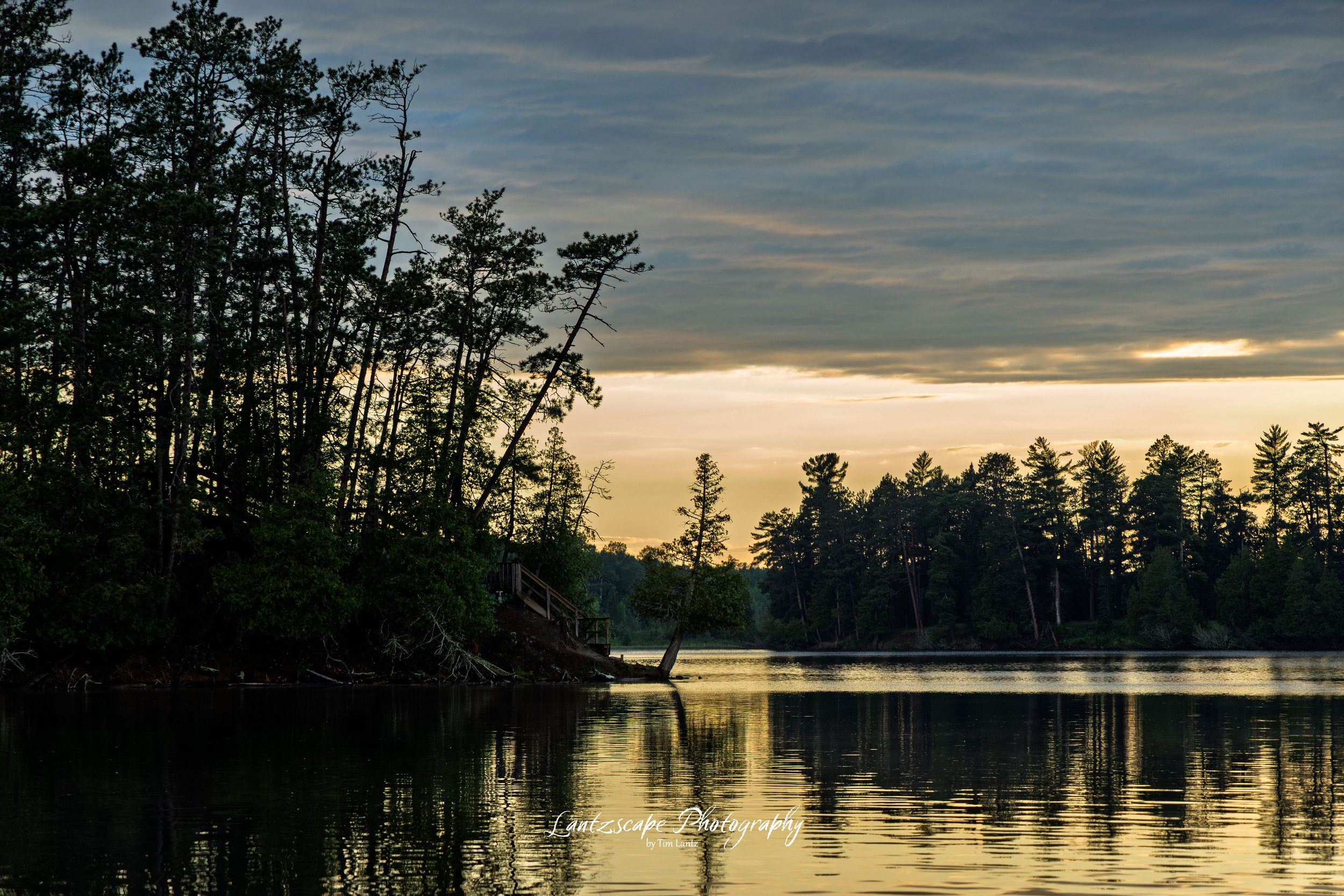A serene landscape of a lake surrounded by trees at sunset, with a partly cloudy sky and calm waters reflecting the scenery.