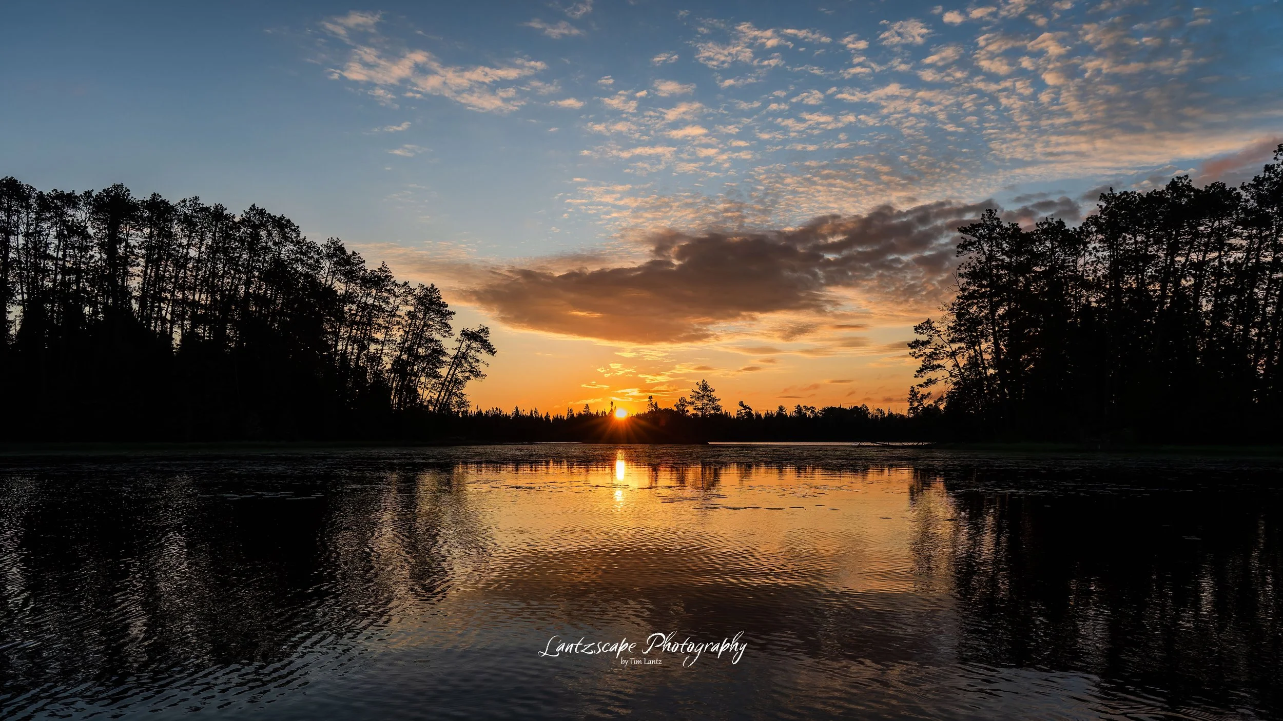 Sunset over a lake with silhouetted trees on both sides, reflecting in the water. The sky has scattered clouds illuminated by the setting sun.