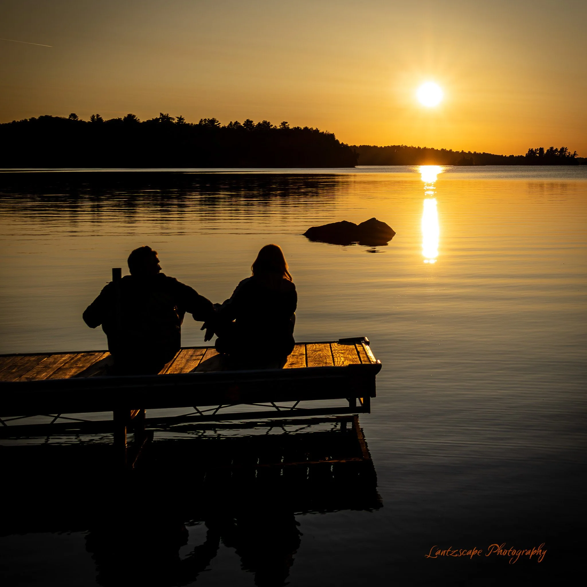 Silhouette of a couple sitting on a dock by a lake at sunset with the sun reflecting on the water, trees in the background.
