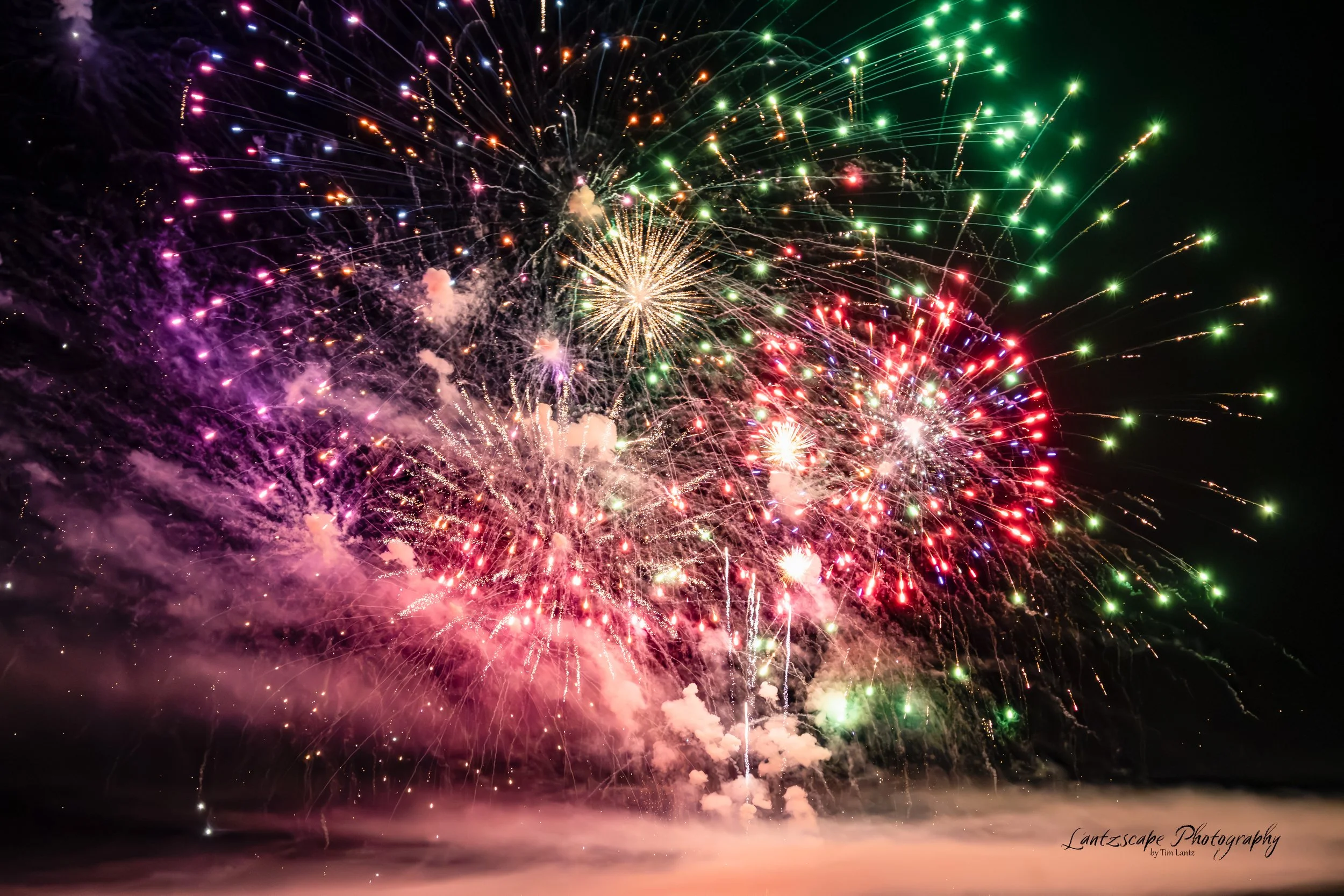 Colorful fireworks explode in the night sky, illuminating the darkness with vibrant reds, greens, purples, and whites.