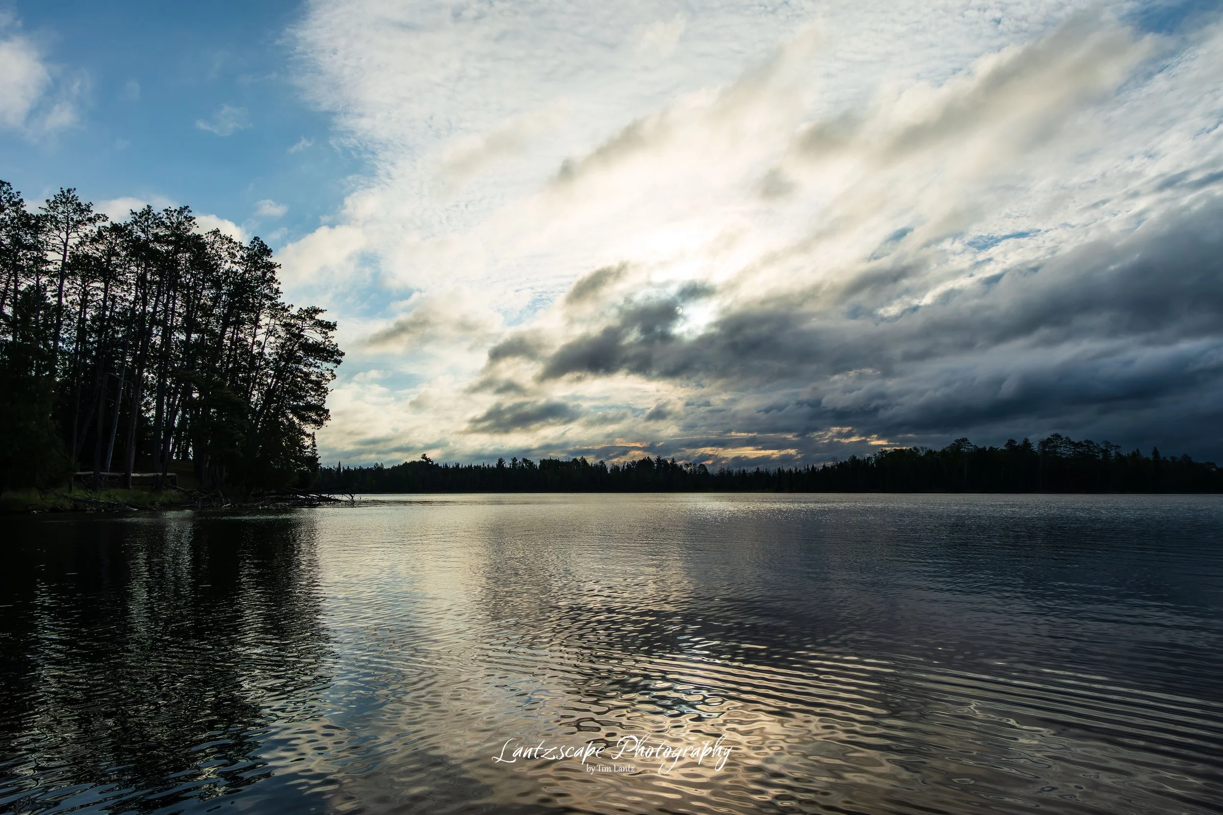 A lake with calm water reflects the partly cloudy sky above. Tall trees line the lakeshore on the left, and distant treetops are visible across the water. The sky is filled with fluffy white clouds and some darker clouds, suggesting a potential sunset or sunrise.