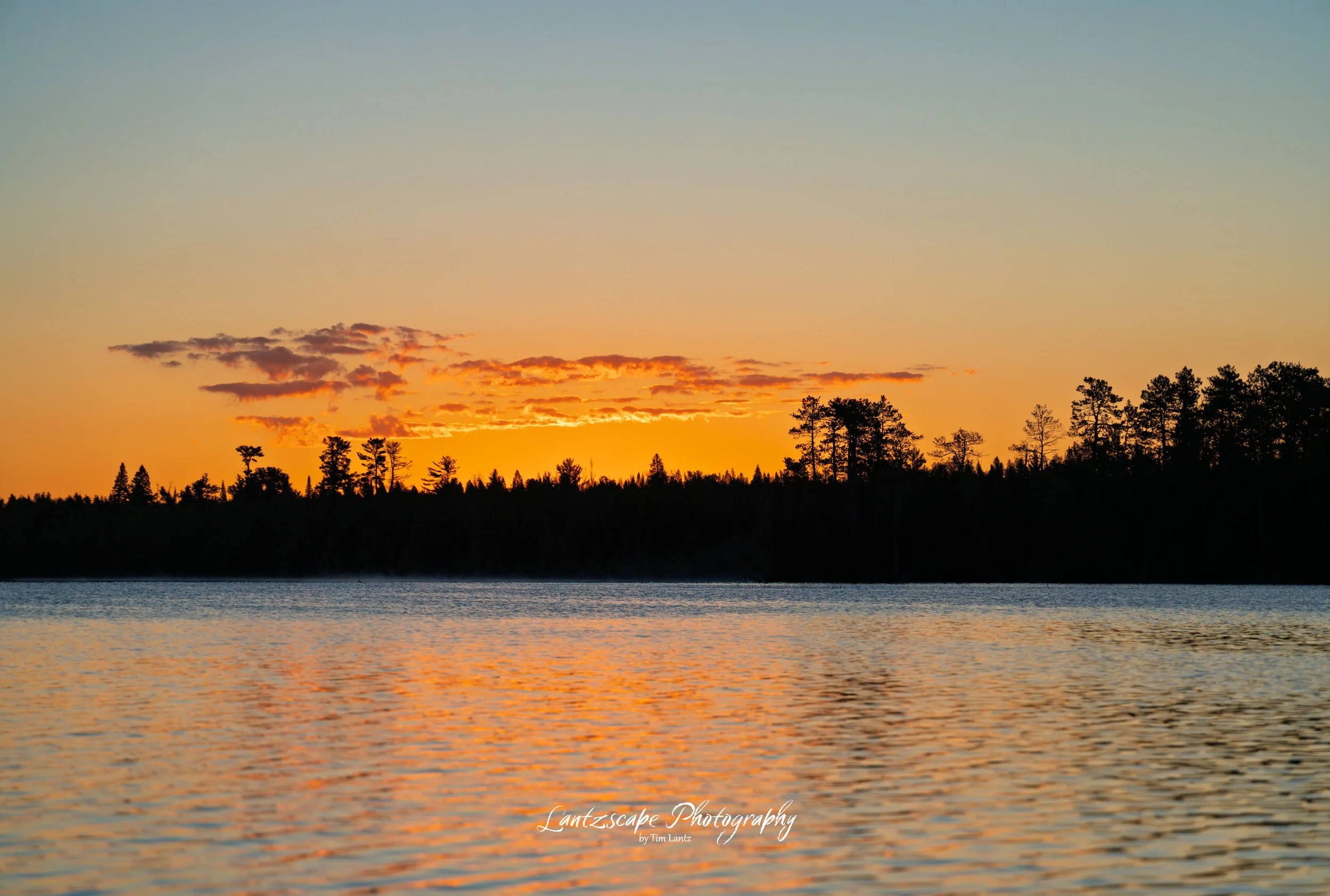 Sunset over a body of water with a tree-lined horizon and scattered clouds in the sky.