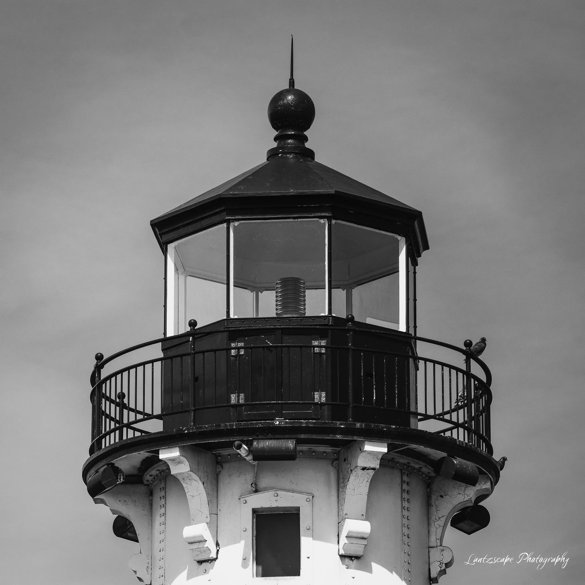 Black and white photo of a lighthouse with a balcony and glass enclosure at the top, against a cloudy sky.