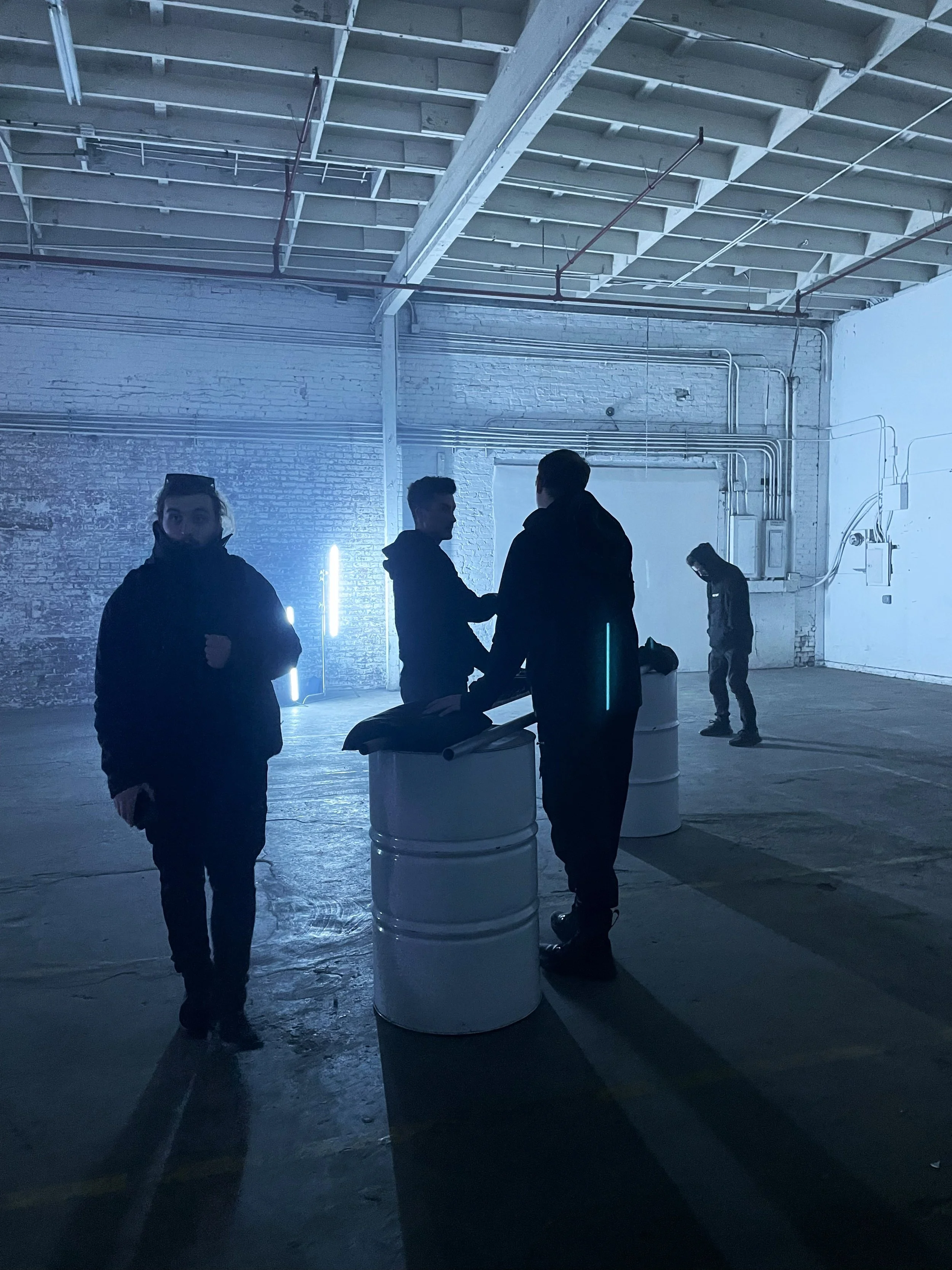 Four people in a dimly lit industrial warehouse with exposed brick walls and pipes. Three are gathered around a table made from barrels, talking, while one stands to the side looking down.