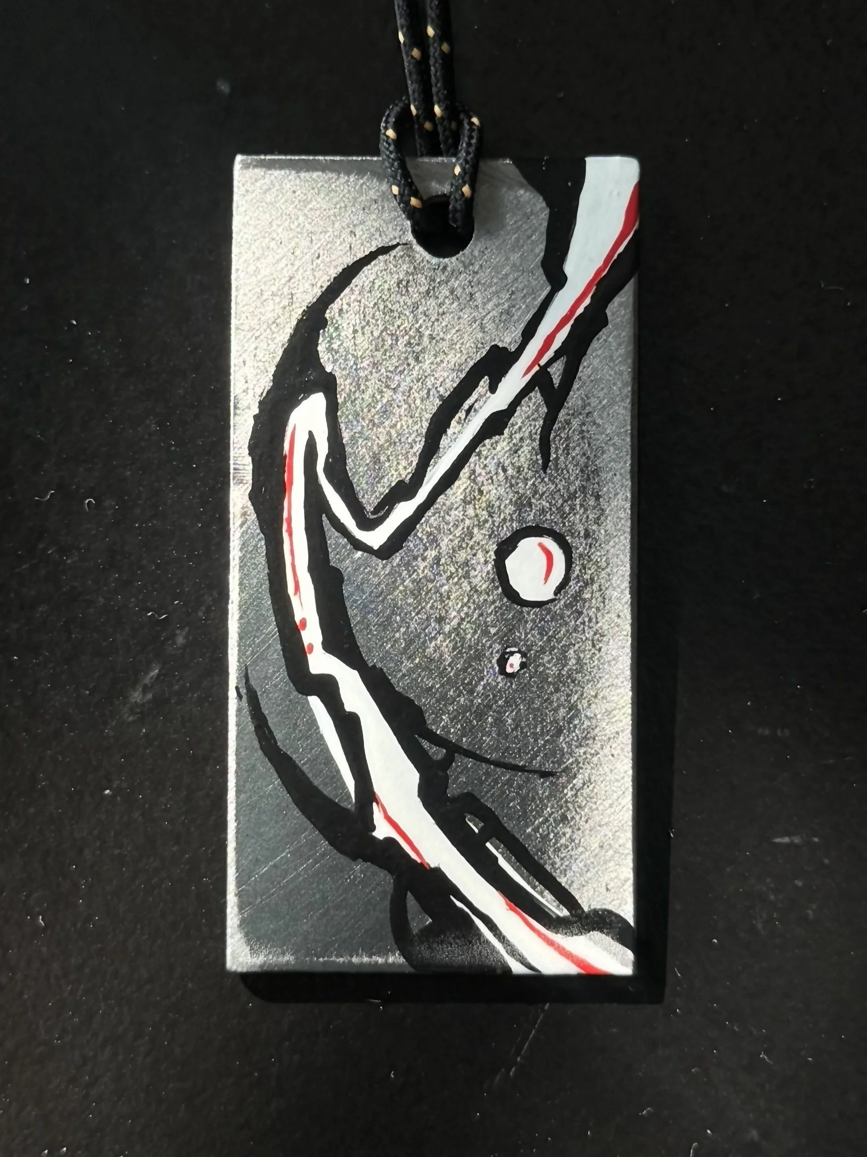 A metal rectangular tag with a black, white, and red graffiti-style design of a baseball player swinging a bat, attached to a black cord.