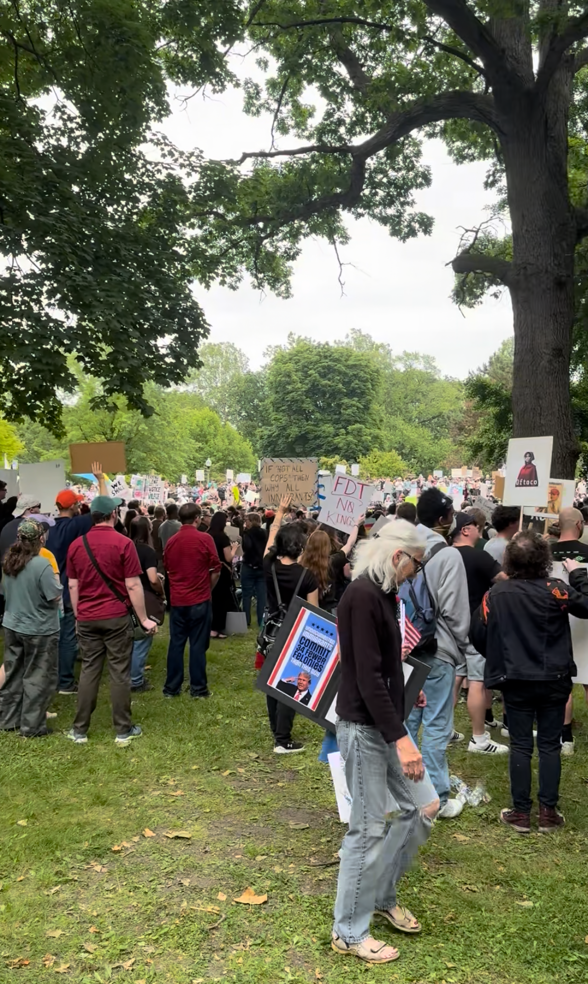 A large crowd of people gathered in a park during a protest or rally, holding various signs with messages about social and political issues. The scene is outdoors, with trees and greenery surrounding the participants.