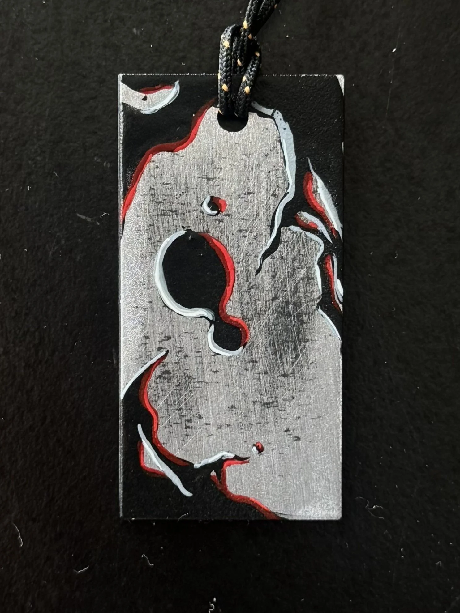 A metallic pendant featuring an outline of a woman's face with stylized red and white accents on a black background, hanging from a black cord.