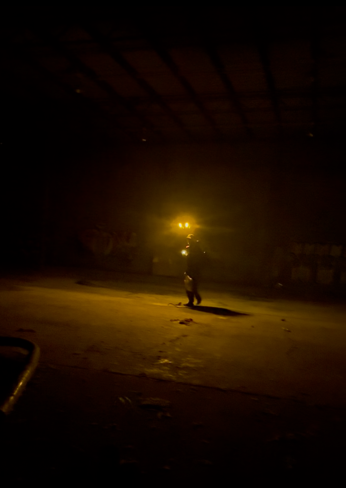 A night scene with a person standing on a skateboard under yellow streetlights in an empty parking lot.