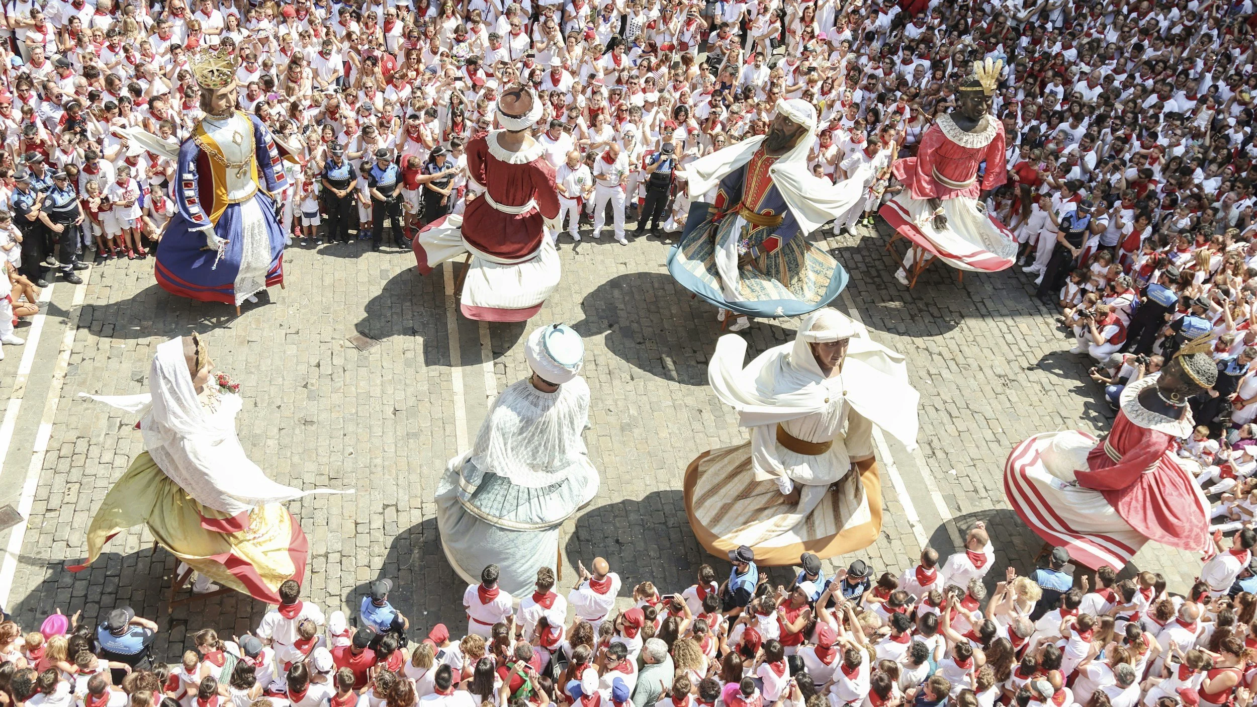 People in traditional costumes perform a dance or parade amid a crowd, with some dressed as kings or queens, in an outdoor celebration or festival.