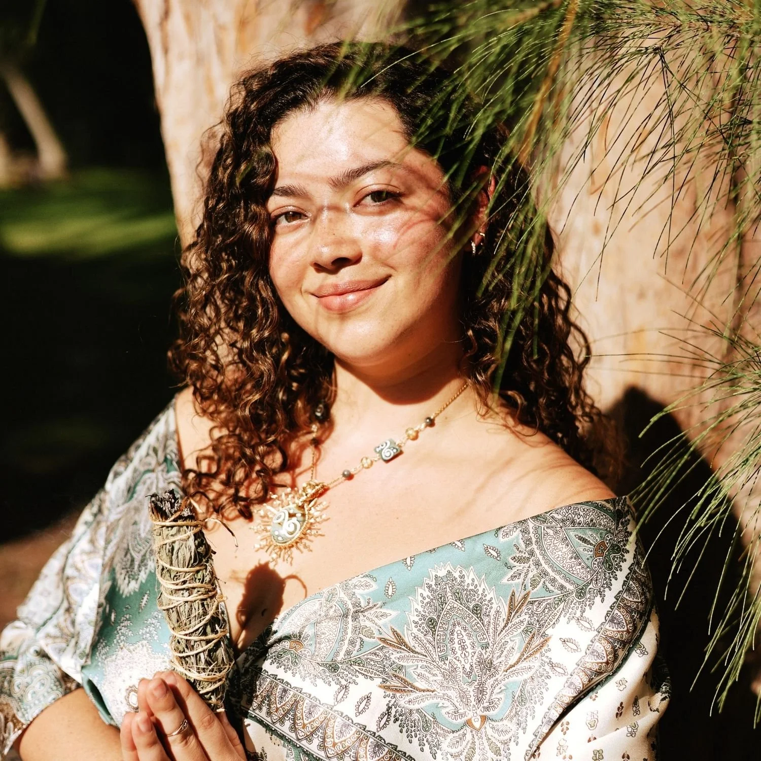 A woman with curly hair smiling outdoors, holding a bundle of sage, wearing a paisley patterned blouse and a necklace with spiritual symbols, partially obscured by a branch with green leaves.