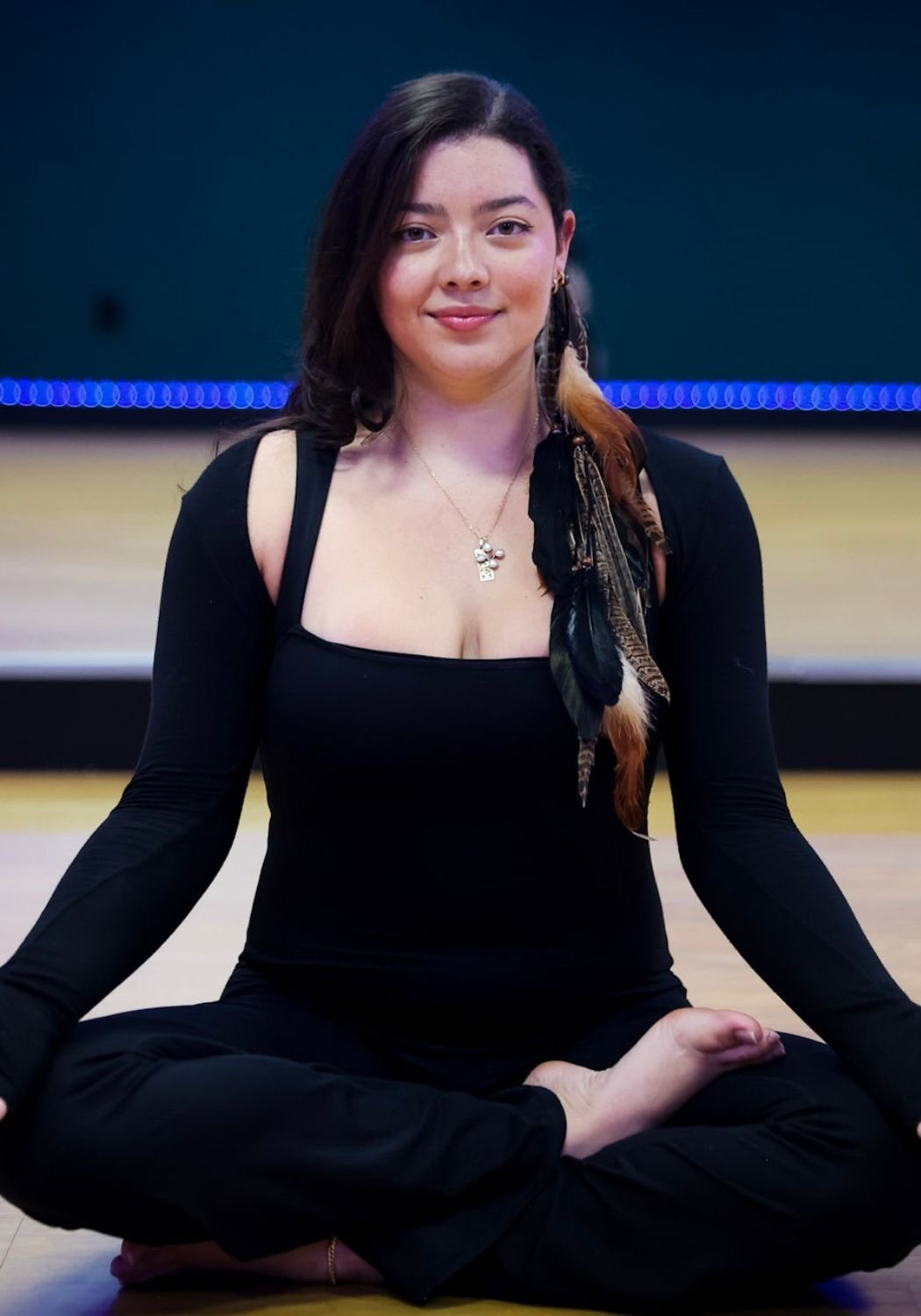 A woman with dark hair, wearing a black outfit and sitting cross-legged on the floor, practicing yoga or meditation in a studio with a wooden floor and blue LED lights in the background.