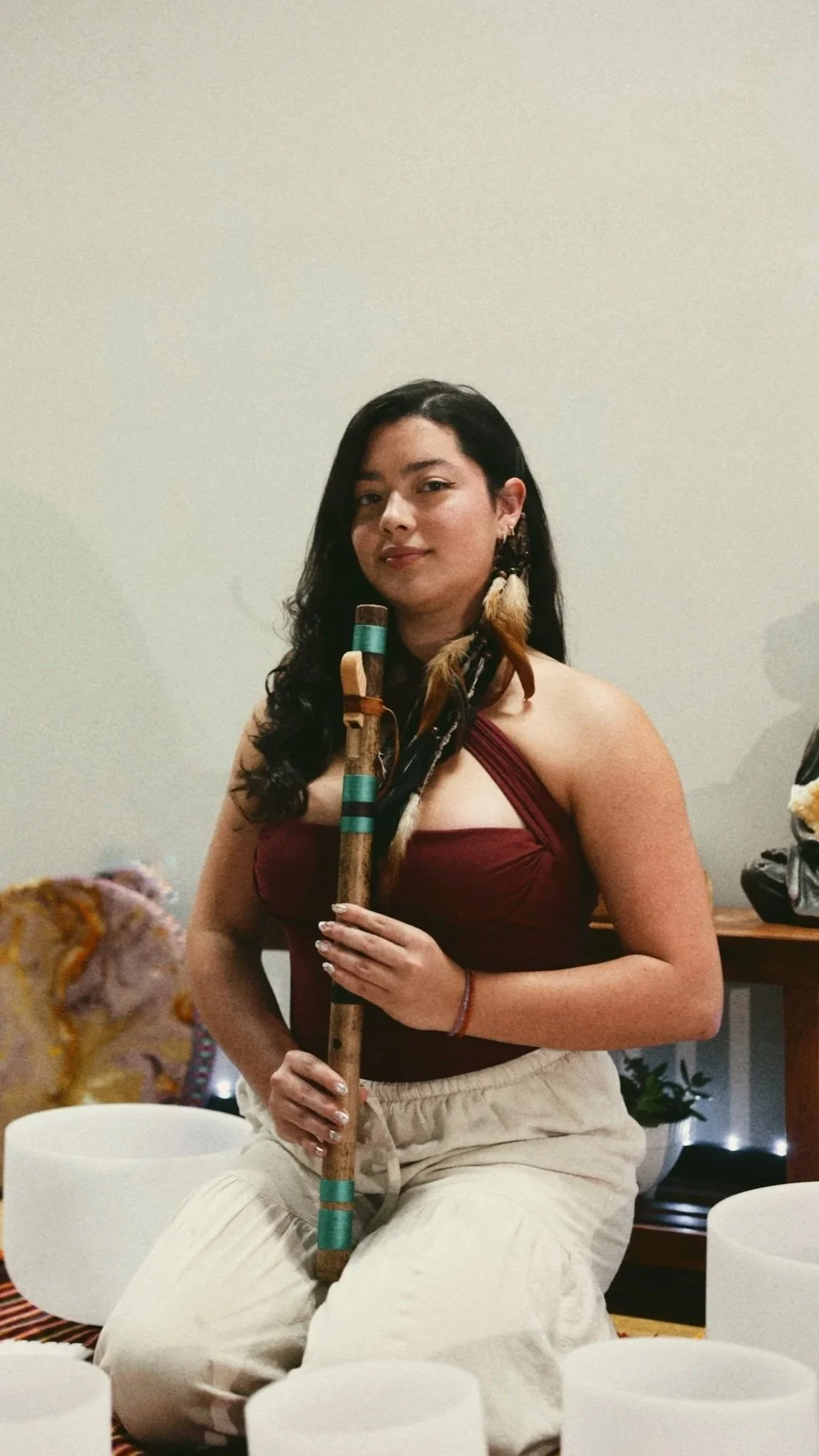 A woman with long dark hair, wearing a maroon top and beige pants, sitting on the floor, holding a wooden flute, surrounded by musical instruments.