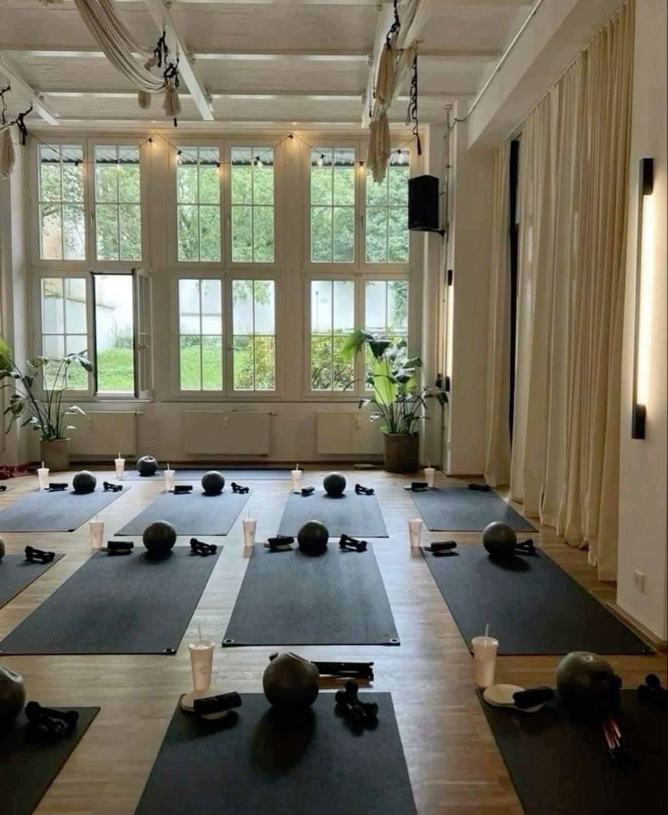 An indoor yoga or fitness class setup with black mats, black medicine balls, small water bottles, and resistance bands on each mat. The room has large windows, potted plants, and curtain-covered walls, creating a bright and airy environment.