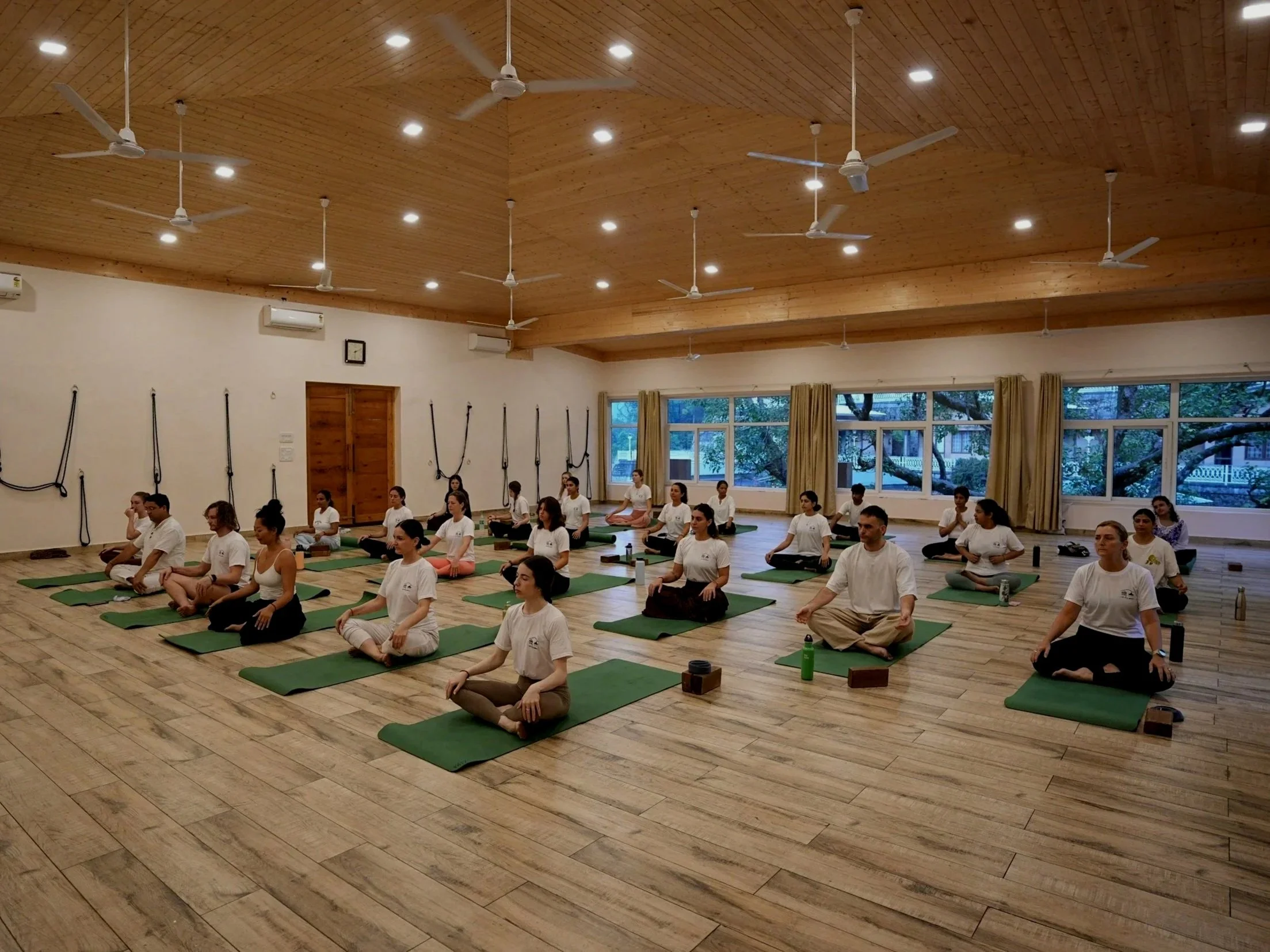 A group of people practicing yoga in a spacious studio with wooden floors and ceiling, sitting on green mats in a cross-legged meditation pose. Large windows with curtains allow natural light into the room.