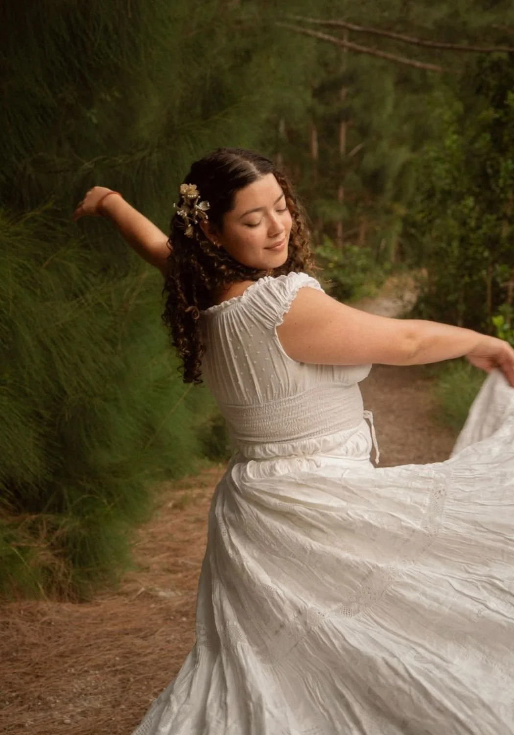 A woman in a white dress is twirling on a forest path with greenery around her, her eyes closed and a peaceful expression on her face.