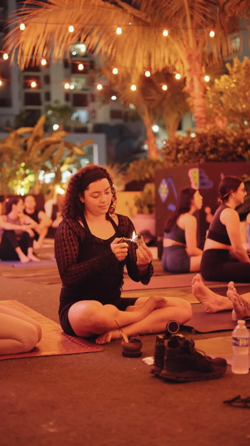 A woman sitting cross-legged on a yoga mat outdoors at night, holding a lit match or fire starter, preparing to ignite incense or a similar item, with other people practicing yoga or meditation in the background under warm string lights and palm trees.