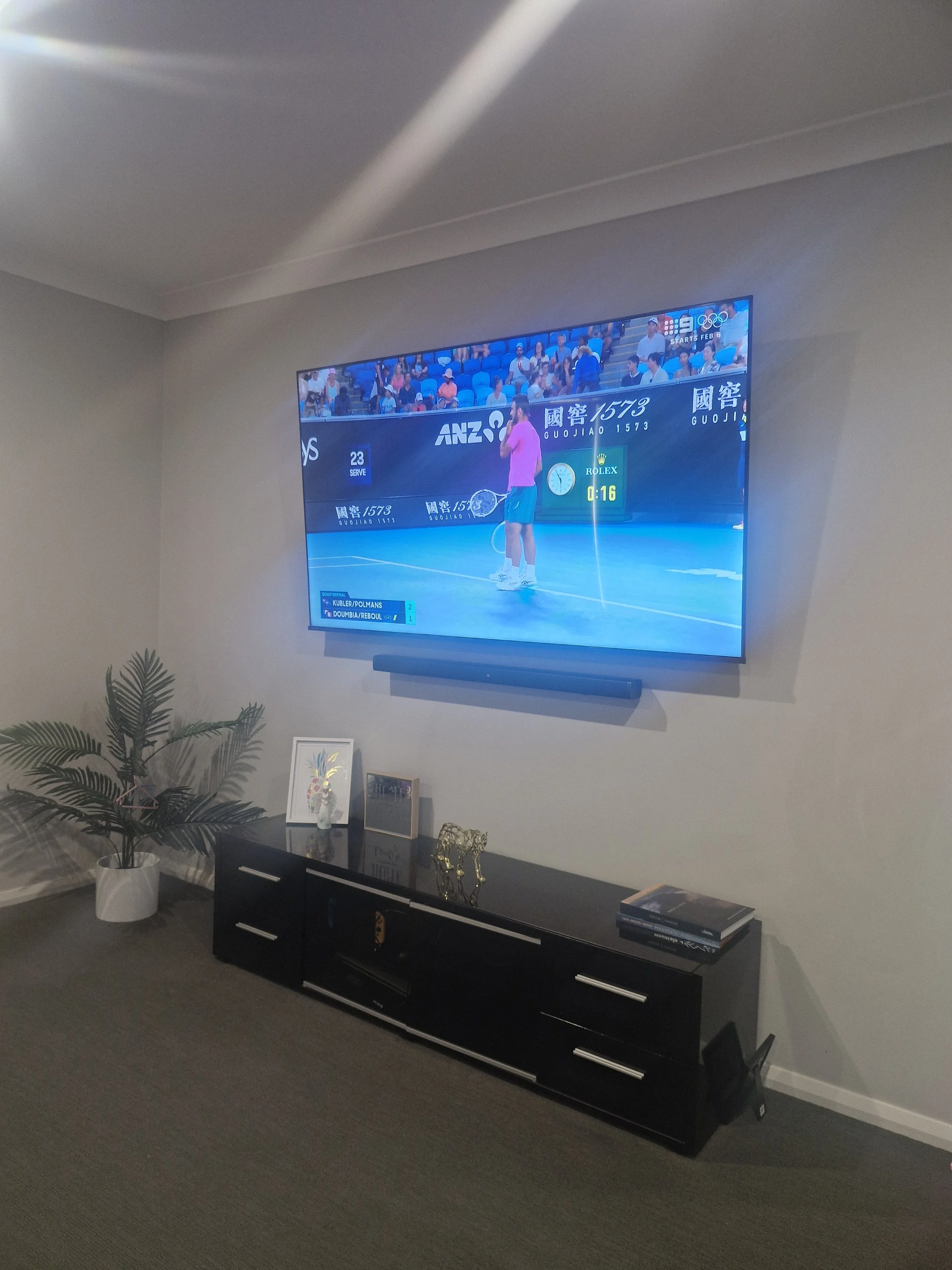 A living room with a wall-mounted flat-screen TV showing a tennis match. Below the TV, there's a black console table with decorative items and books.