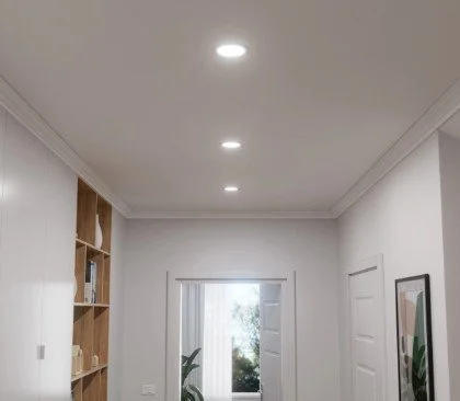 Interior of a modern room with white walls, ceiling lights, a wooden bookshelf on the left, a door on the right, and a window with a view of trees outside.