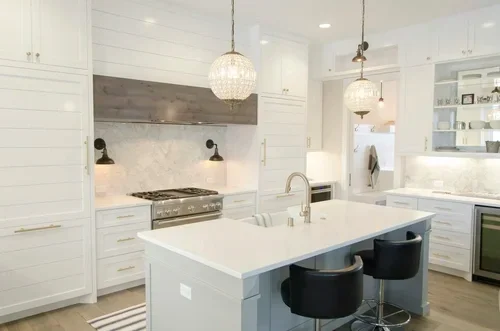 Modern white kitchen with island and black bar stools, pendant lighting, and built-in shelves with dishes.