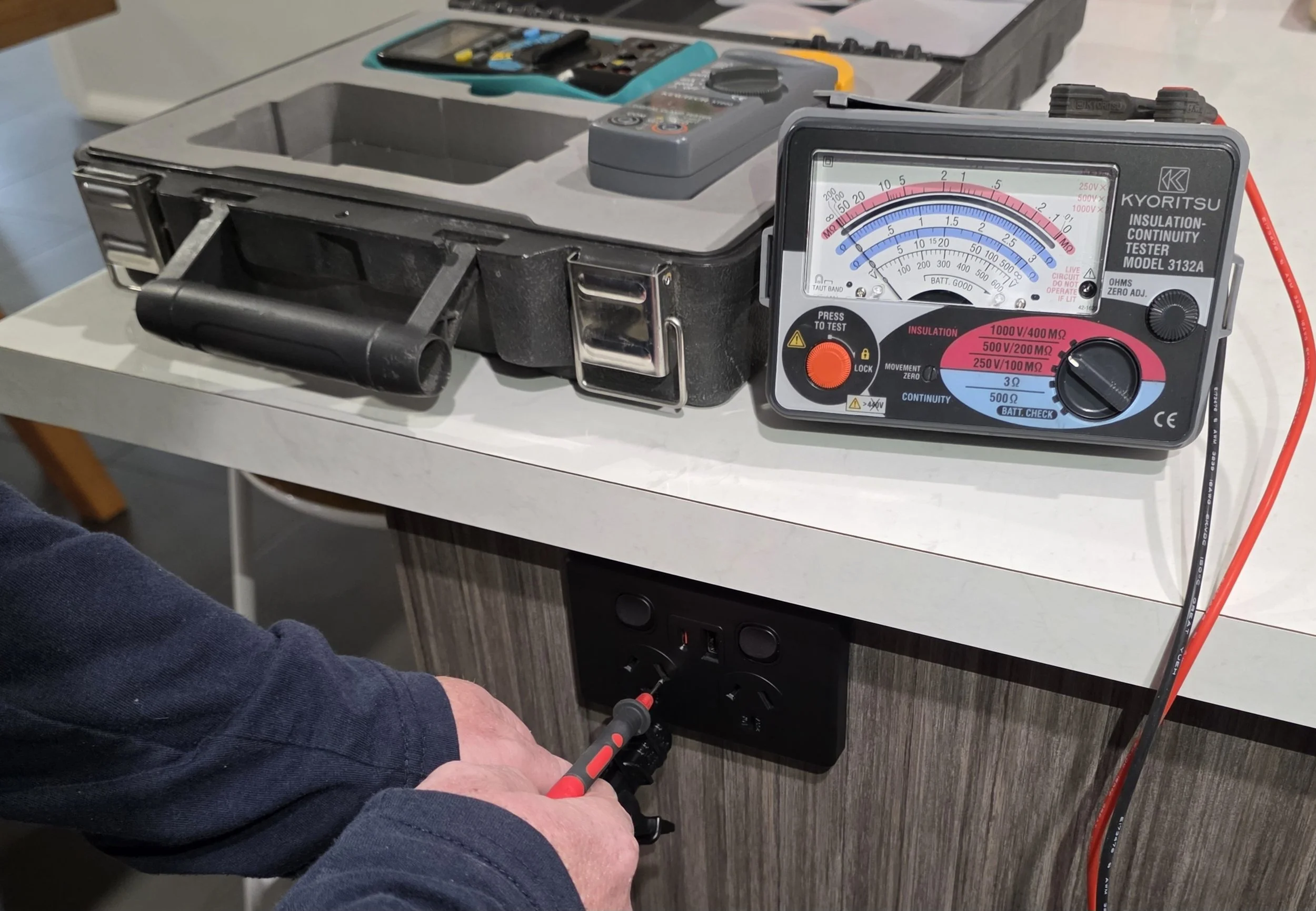A person using a digital multimeter to test an electrical outlet, with a test box and tools on a white table.
