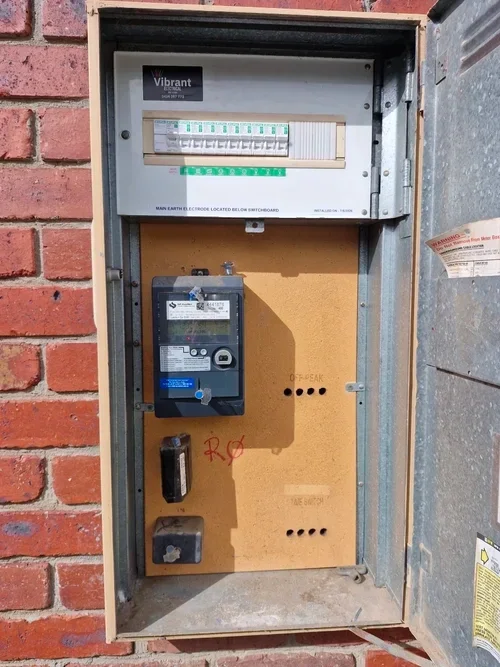 An open electrical meter box mounted on a wall, containing a digital electricity meter and a circuit breaker panel with multiple switches.