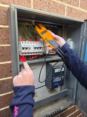 Person using a digital multimeter to test electrical connections inside an outdoor electrical panel mounted on a brick wall.