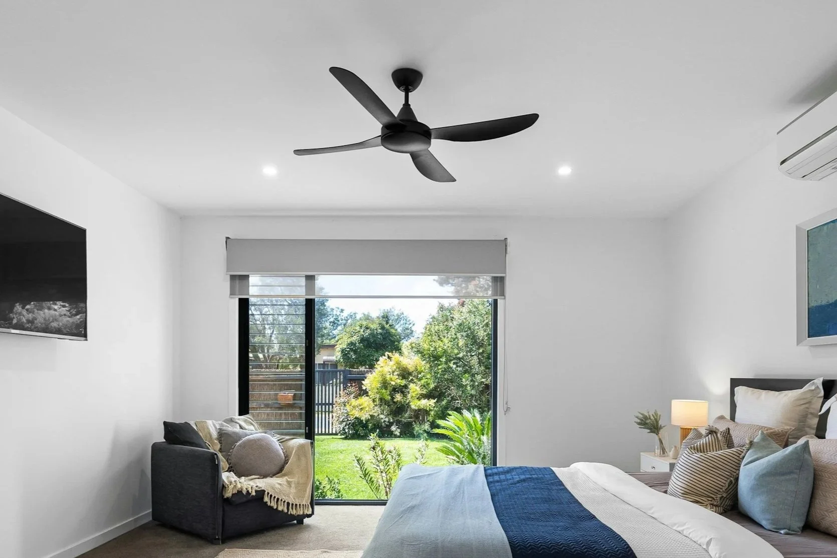 Modern bedroom with white walls, a large window with a view of greenery, a bed with striped and solid pillows, a bedside table with a lamp and plant, a wall-mounted TV, a ceiling fan, and a blanket draped over a small armchair near the window.