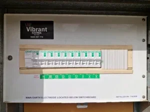Electrical switchboard with main earth electrode label beneath, containing multiple green circuit breakers and a black sticker with 'Vibrant Electrical' logo and contact number.