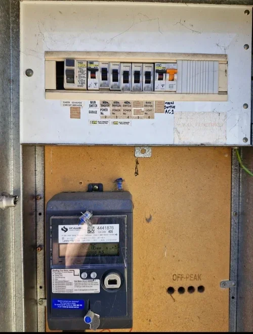 An electrical breaker panel with circuit breakers and labels in the upper section, a digital utility meter below, and a beige wall with a small black hole labeled "OFF-PEAK" on the right side.