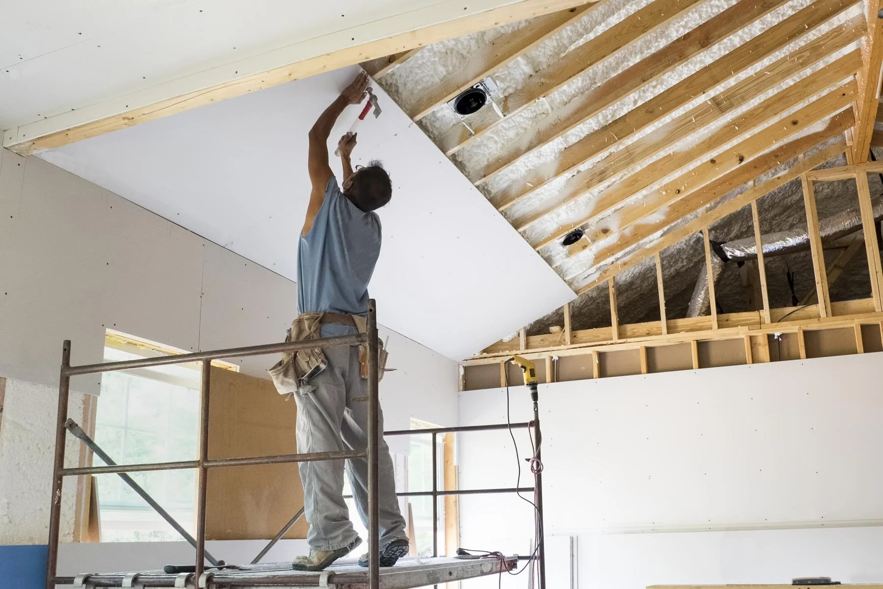 Construction worker standing on scaffolding installing drywall on a sloped ceiling in a building under construction.