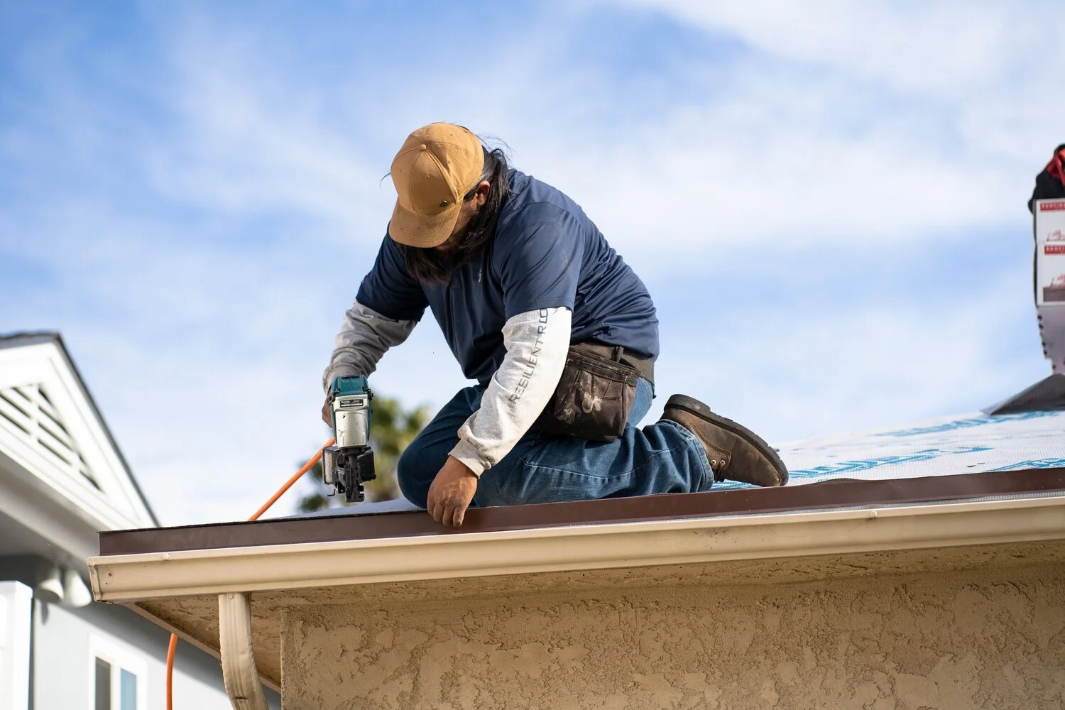 A construction worker kneels on a roof, using a power tool to work on the edge of the roof's metal trim.
