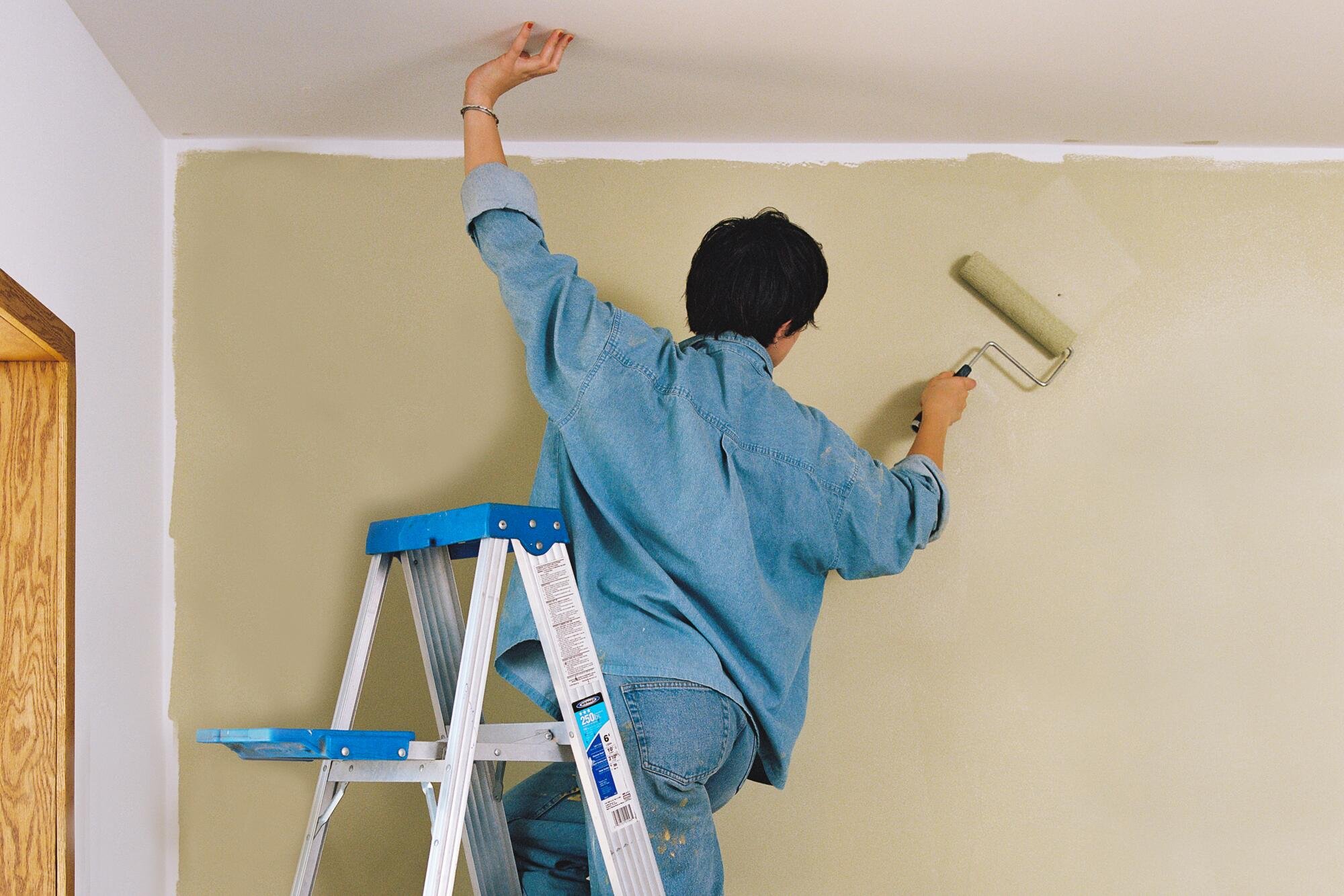 Person painting a wall cream color with a paint roller while standing on a step ladder.