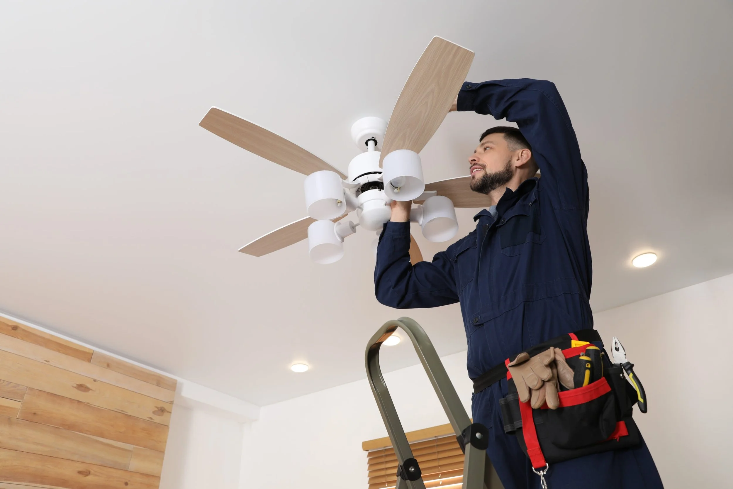 Man installing a ceiling fan while standing on a ladder in a room with white walls and wooden accents.