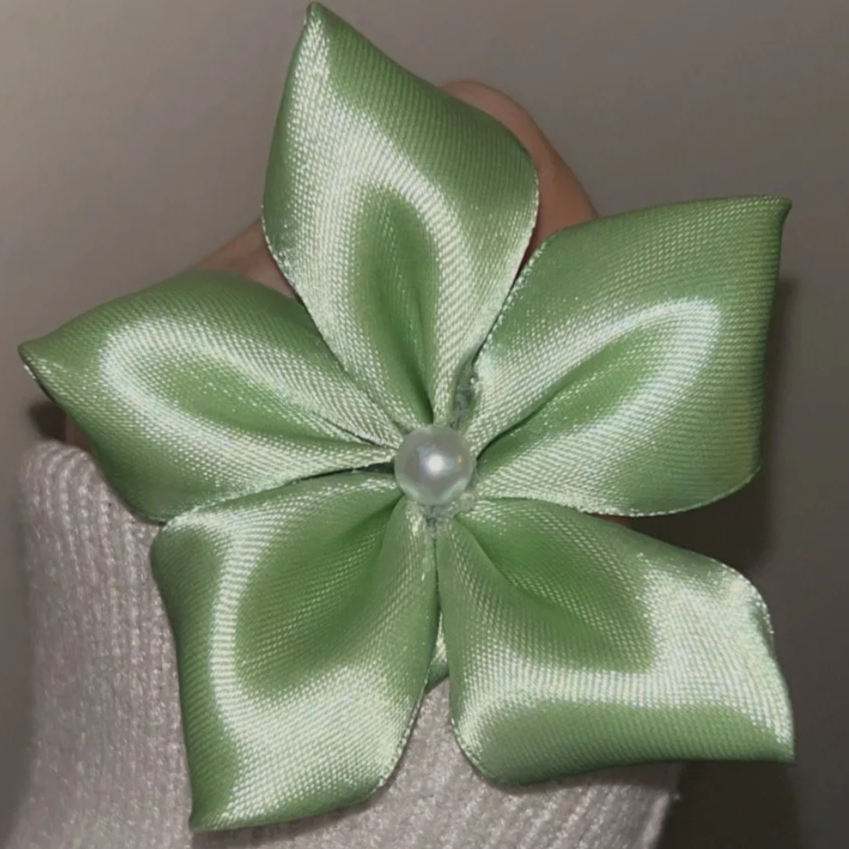 Green satin ribbon flower with a pearl center, attached to a fabric surface.