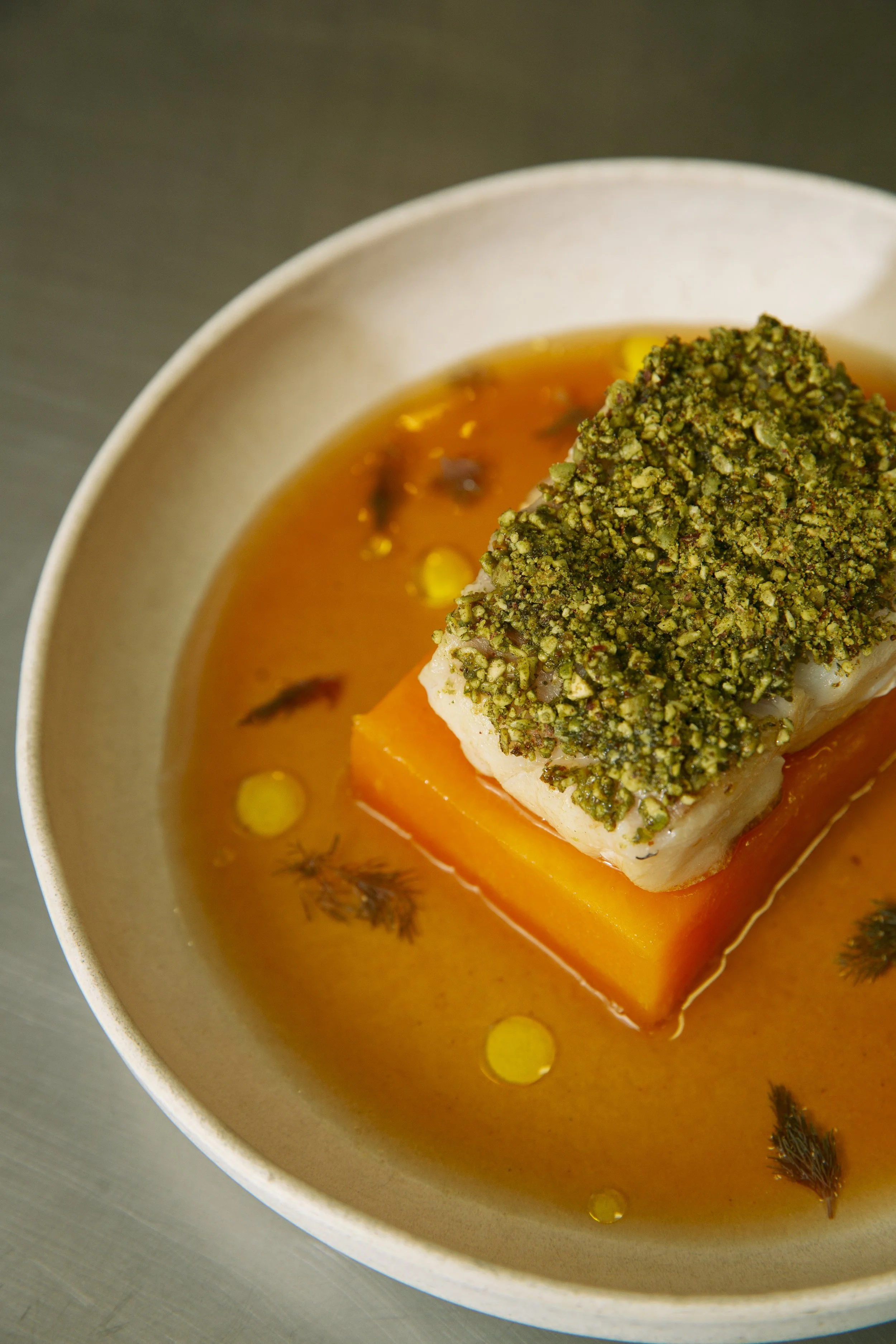 A bowl of fish stew with a layer of broth, containing a fish fillet coated with crushed pistachios, placed on a piece of butternut squash, and garnished with herbs and oil droplets.
