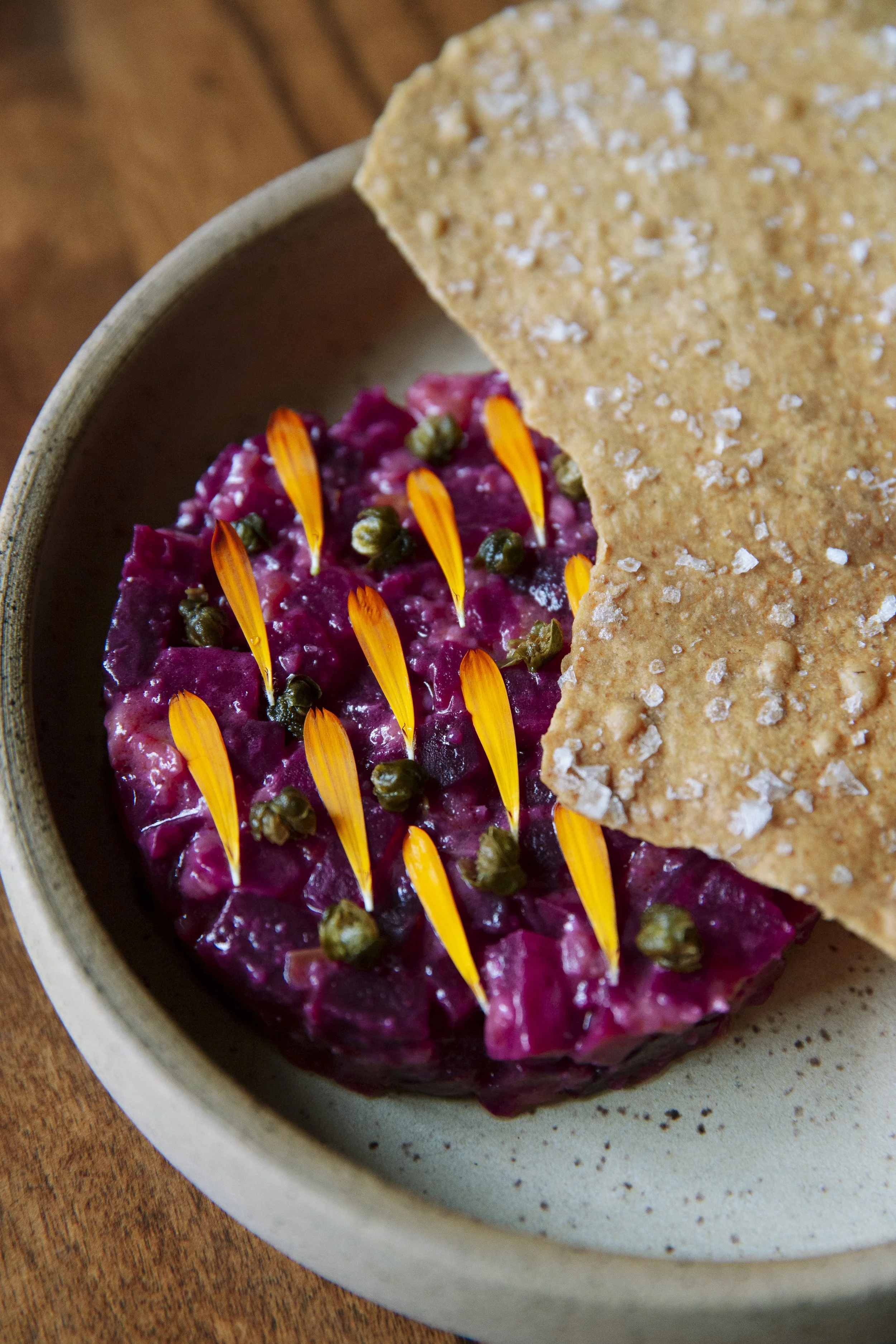 A bowl of beet salad garnished with orange flower petals and capers, with a piece of salt-crusted flatbread on the side.