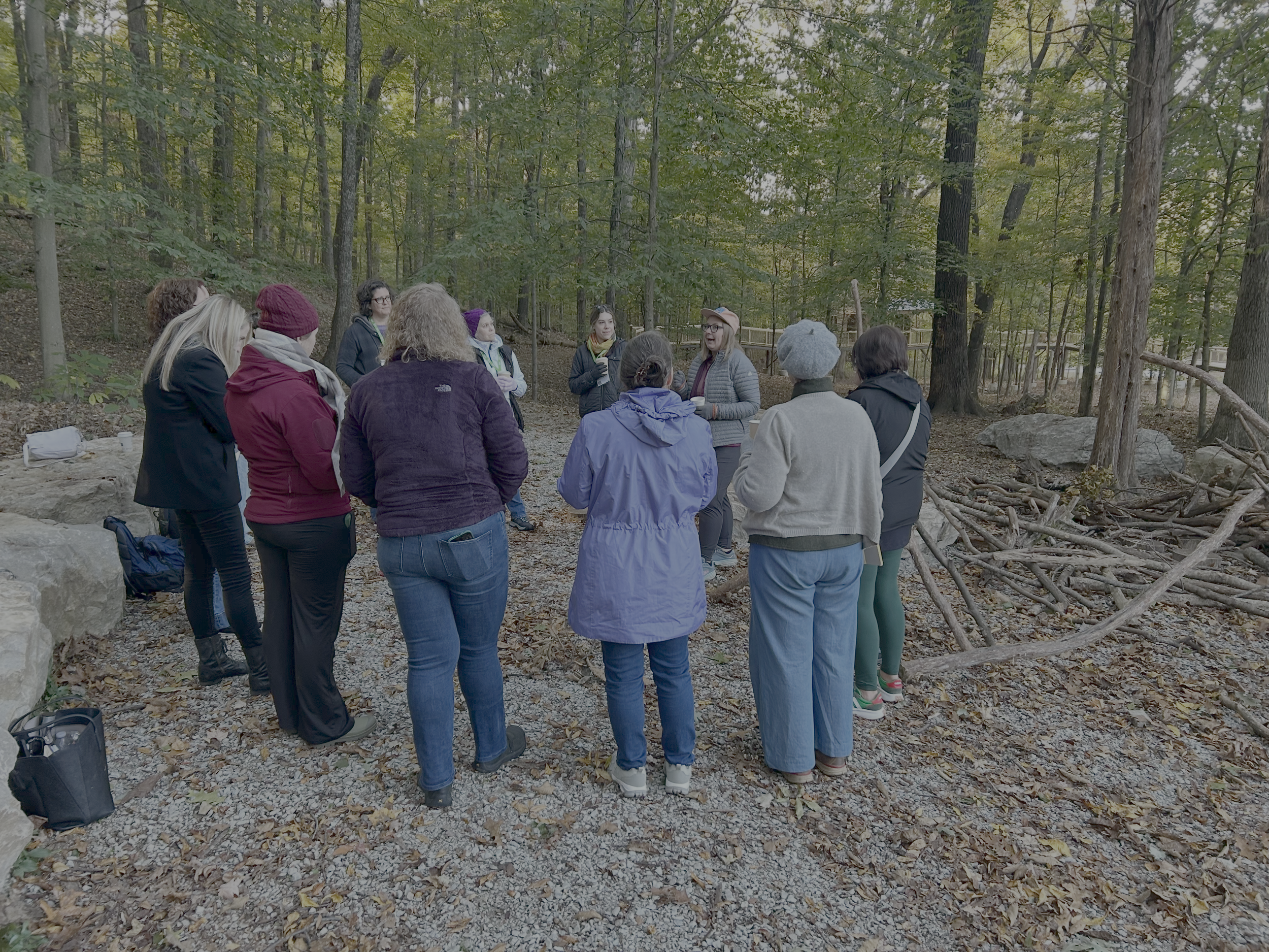 A group of people gathered in a semi-circle outdoors in a wooded area with tall trees and fallen leaves, appears to be listening to a woman speaking.