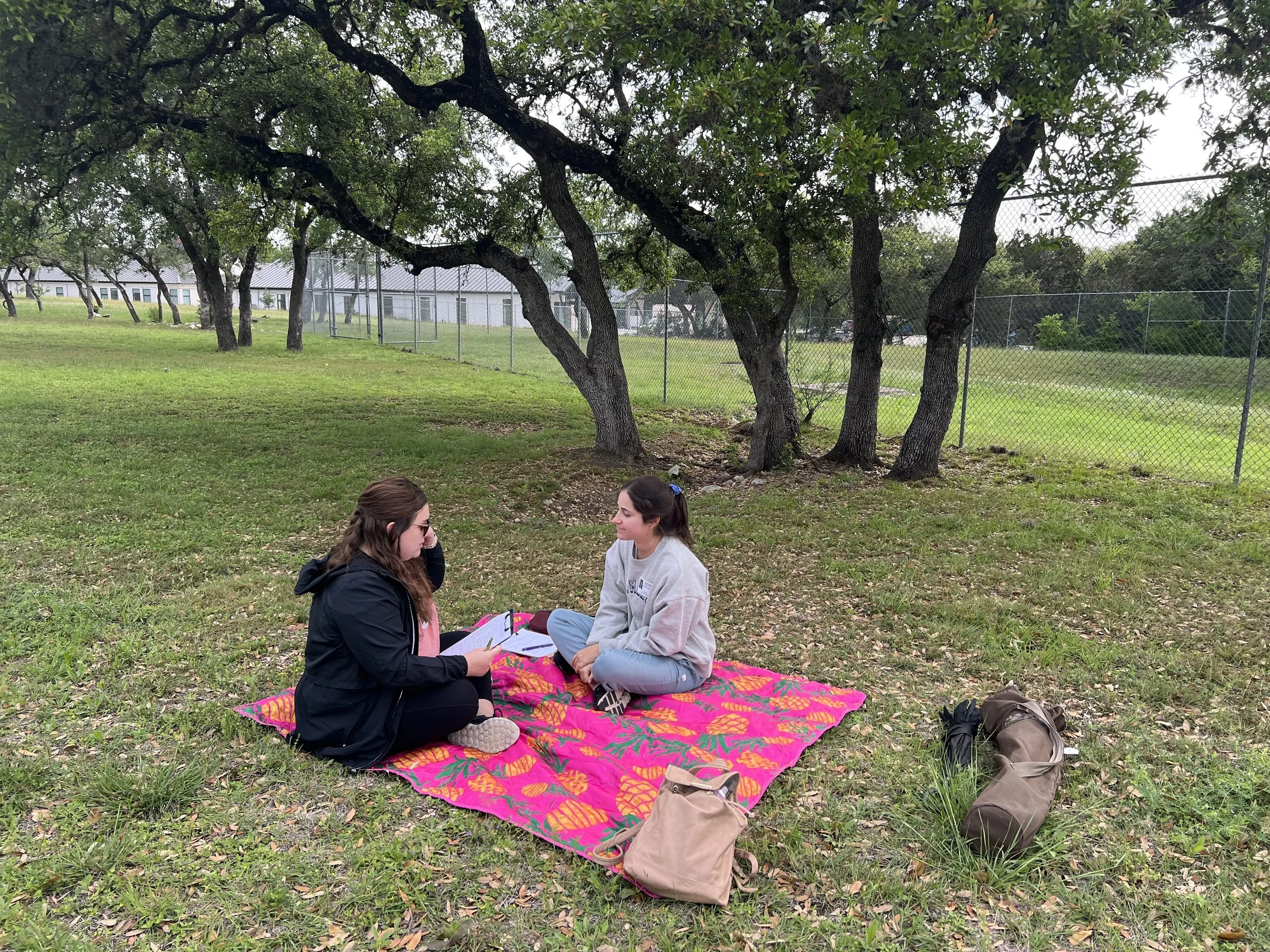 Two women sitting cross-legged on a colorful blanket under trees in a park, engaged in conversation, with backpacks nearby.