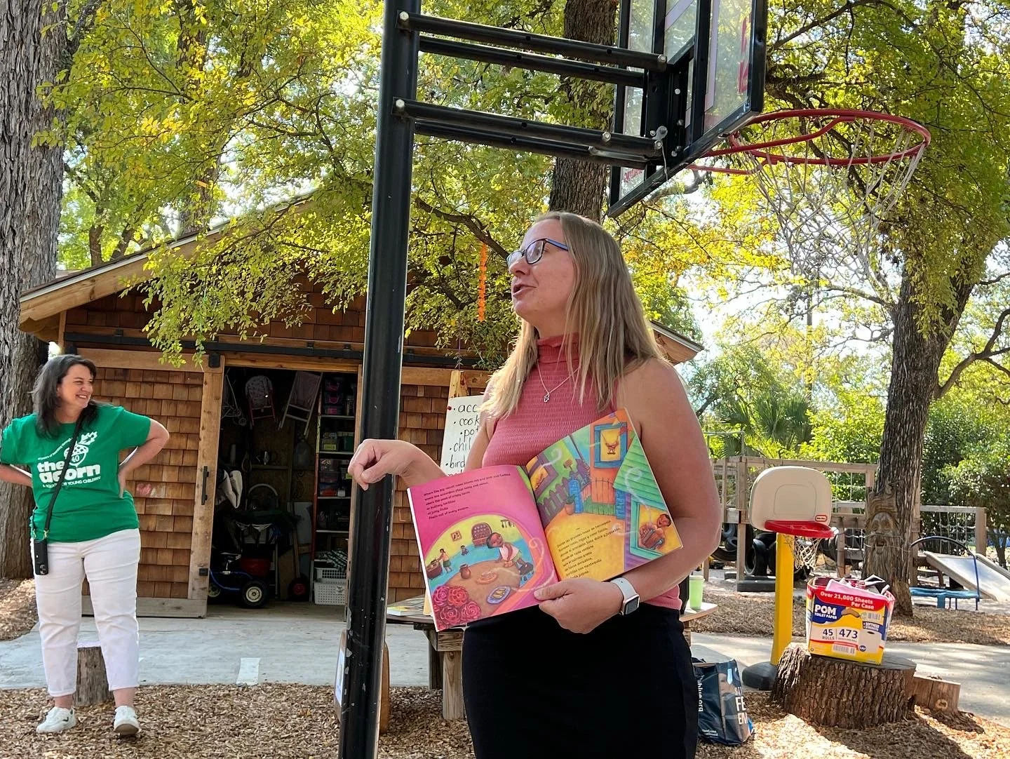 Woman with glasses, pink sleeveless top, and black pants standing outdoors near a basketball hoop, holding a colorful children's book, with another woman in a green shirt smiling in the background, surrounded by trees and outdoor equipment.