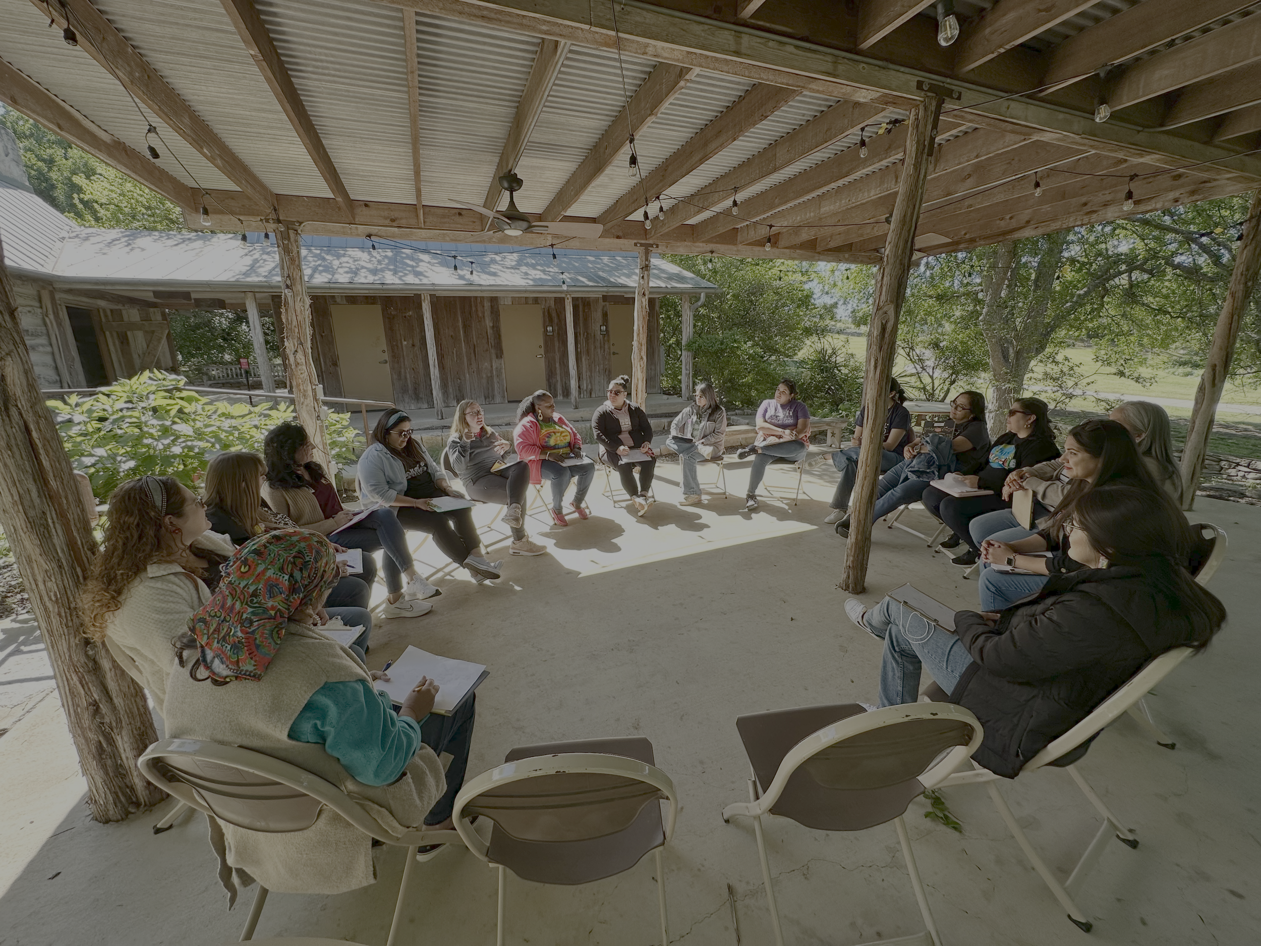 A group of diverse women sitting in a circle outdoors on chairs, engaged in a discussion under a wooden pavilion with string lights, surrounded by trees and greenery.