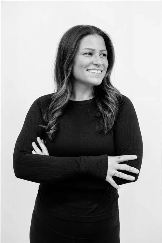 A woman with long, wavy hair smiling and looking to her right, wearing a black long-sleeve shirt, standing against a plain white background.