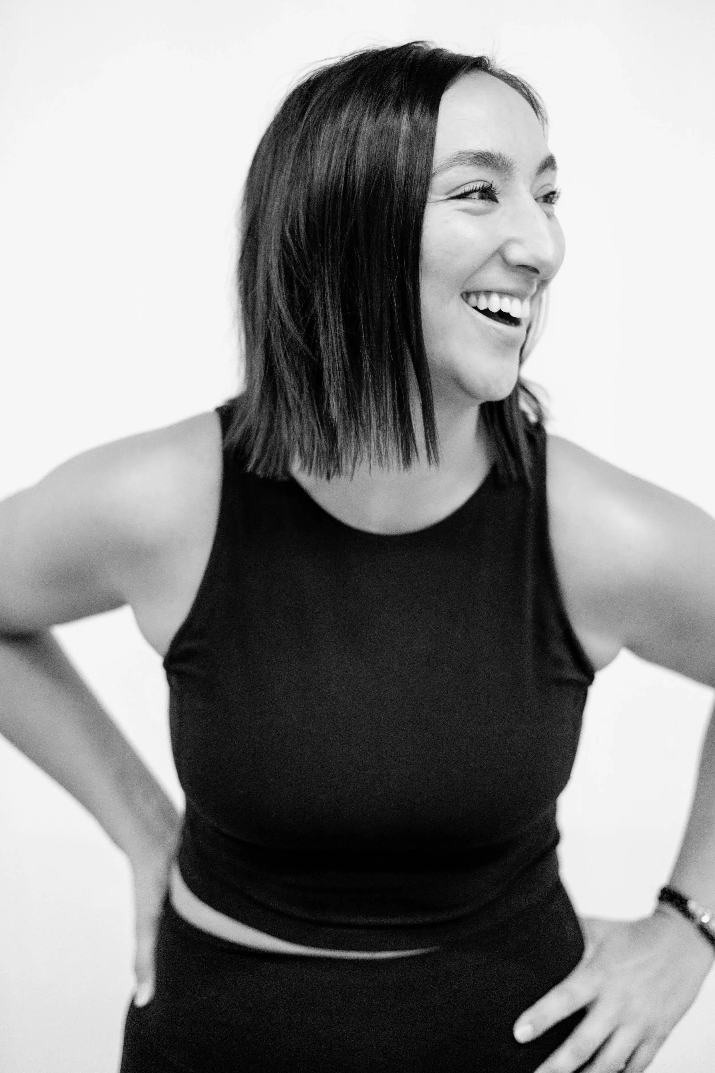 Black and white photo of a woman smiling, with shoulder-length dark hair, wearing a sleeveless dark top, standing with hands on hips, looking to the side.