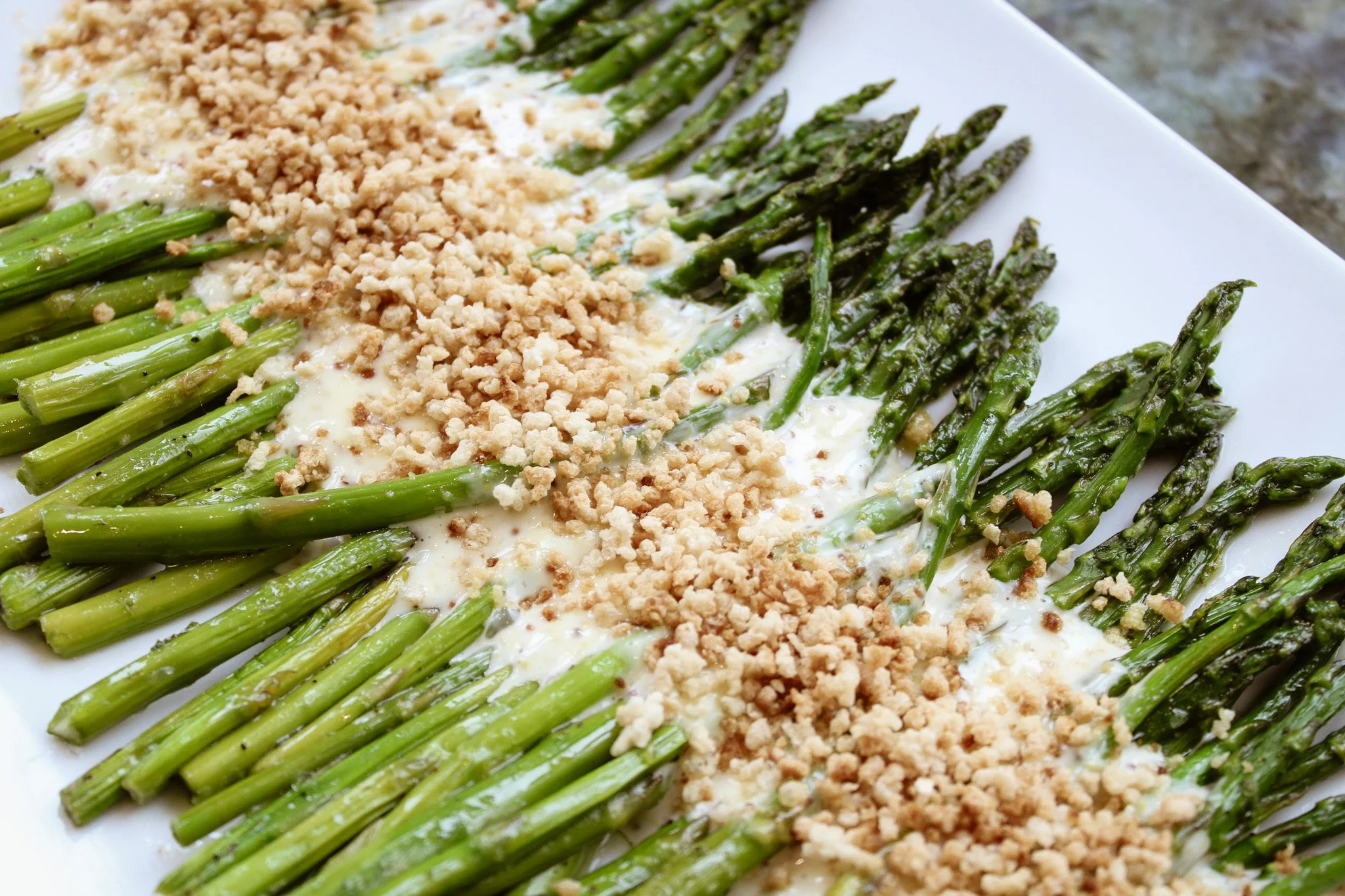 Baked asparagus spears topped with crumbled cheese and breadcrumbs on a white rectangular plate.